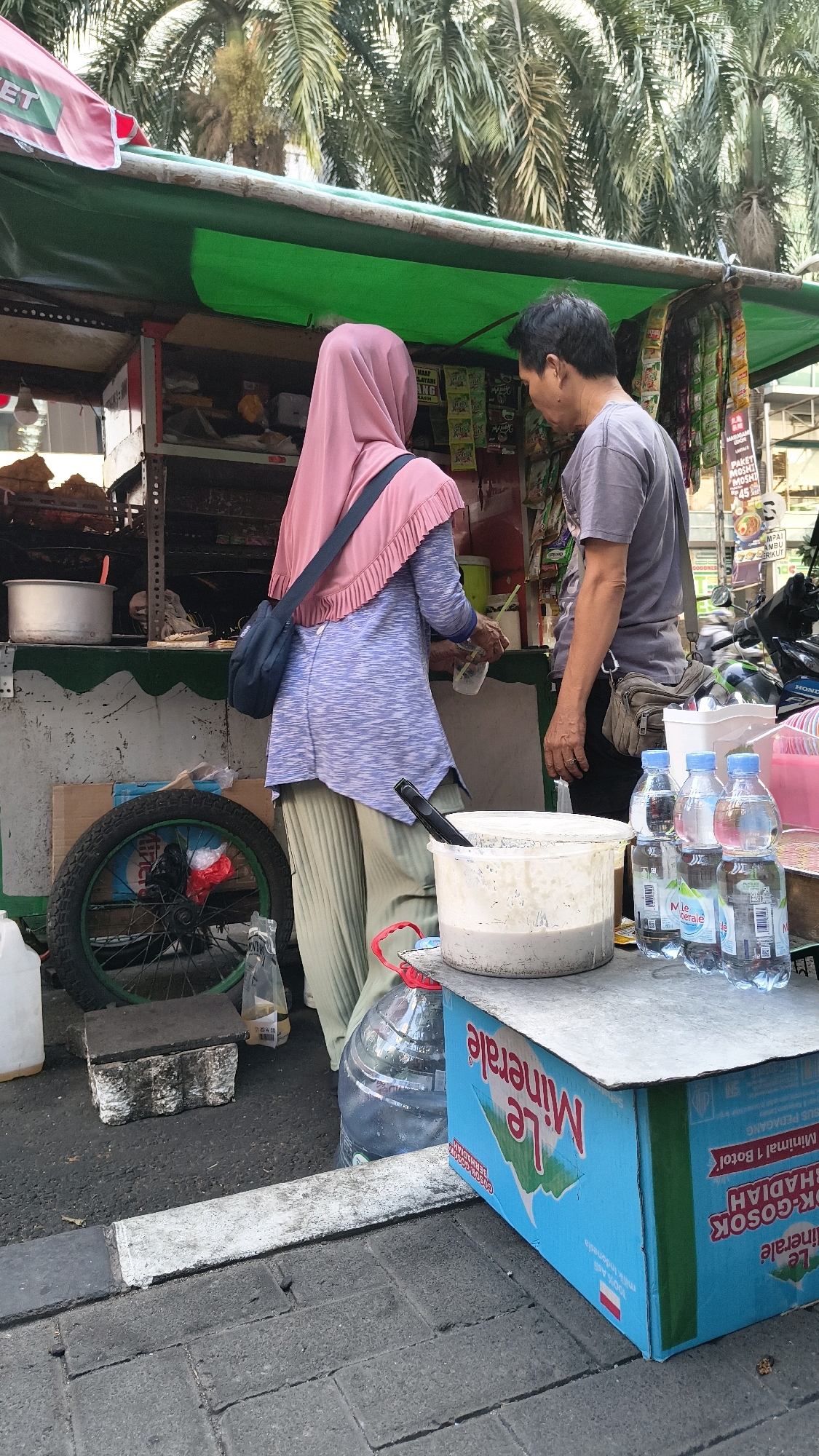 Two individuals, a woman wearing a pink hijab and a purple and white striped shirt with green pants, and a man in a grey t-shirt and dark pants, are standing at a street food stall in South Jakarta, Indonesia. The woman is interacting with the stall, possibly making a purchase, holding a plastic cup and a straw. The man is facing her, looking down towards the stall.
The stall is covered by a green tarp and has various items for sale displayed on shelves. In the foreground, there's a blue cardboard box with the "Le Minerale" logo, several bottles of water, and a large white plastic tub containing a light-colored mixture. A bicycle wheel is visible to the left of the stall. The ground is paved with asphalt and concrete. Palm trees are visible in the background, suggesting an outdoor, possibly urban, environment. The lighting indicates it is daytime.