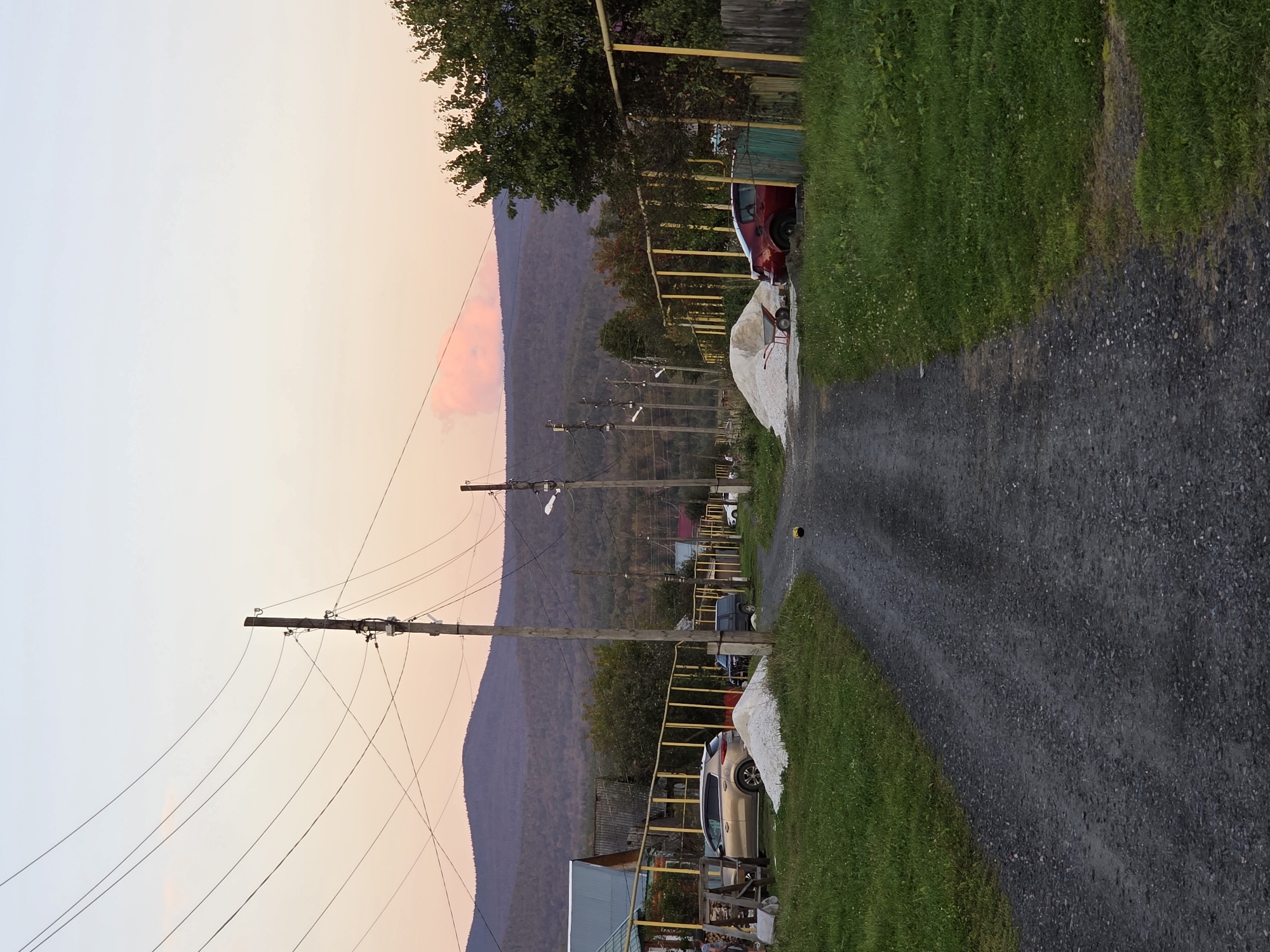 The image depicts a dirt road in a rural area, likely a village, with houses and fences on either side. Power lines stretch across the sky, supported by wooden poles. In the background, a forested mountain range is visible under a pale sky with hints of pink from a sunset. Two cars, one gold and one red, are parked along the road. Piles of white material, possibly gravel or sand, are present near the houses. A wheelbarrow is next to one of the piles. The foreground shows the textured surface of the dirt road and a strip of green grass on the left side. The scene appears to be in Magnitka, Russia.