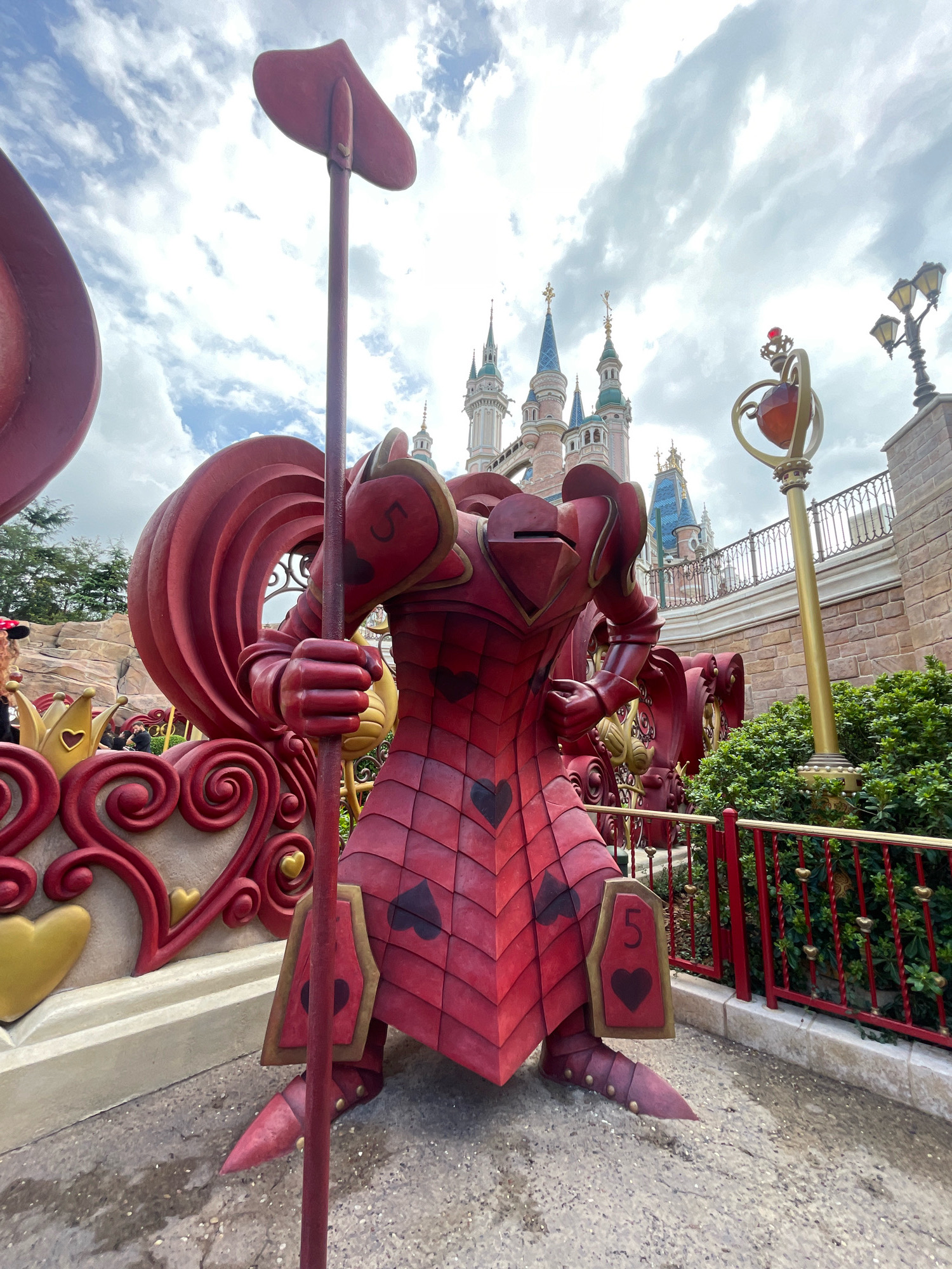 A red, armored statue resembling a playing card is positioned in the foreground, facing right, with a spiked weapon held in its right hand. The statue is adorned with heart symbols and the number "5" on its armor. Behind it, a castle with multiple spires and flags is visible, characteristic of Disneyland. A lamppost with a decorative orb is to the right of the statue. The scene is set against a cloudy sky, with lush green bushes and red railings in the background. This image was taken in Pudong, China.