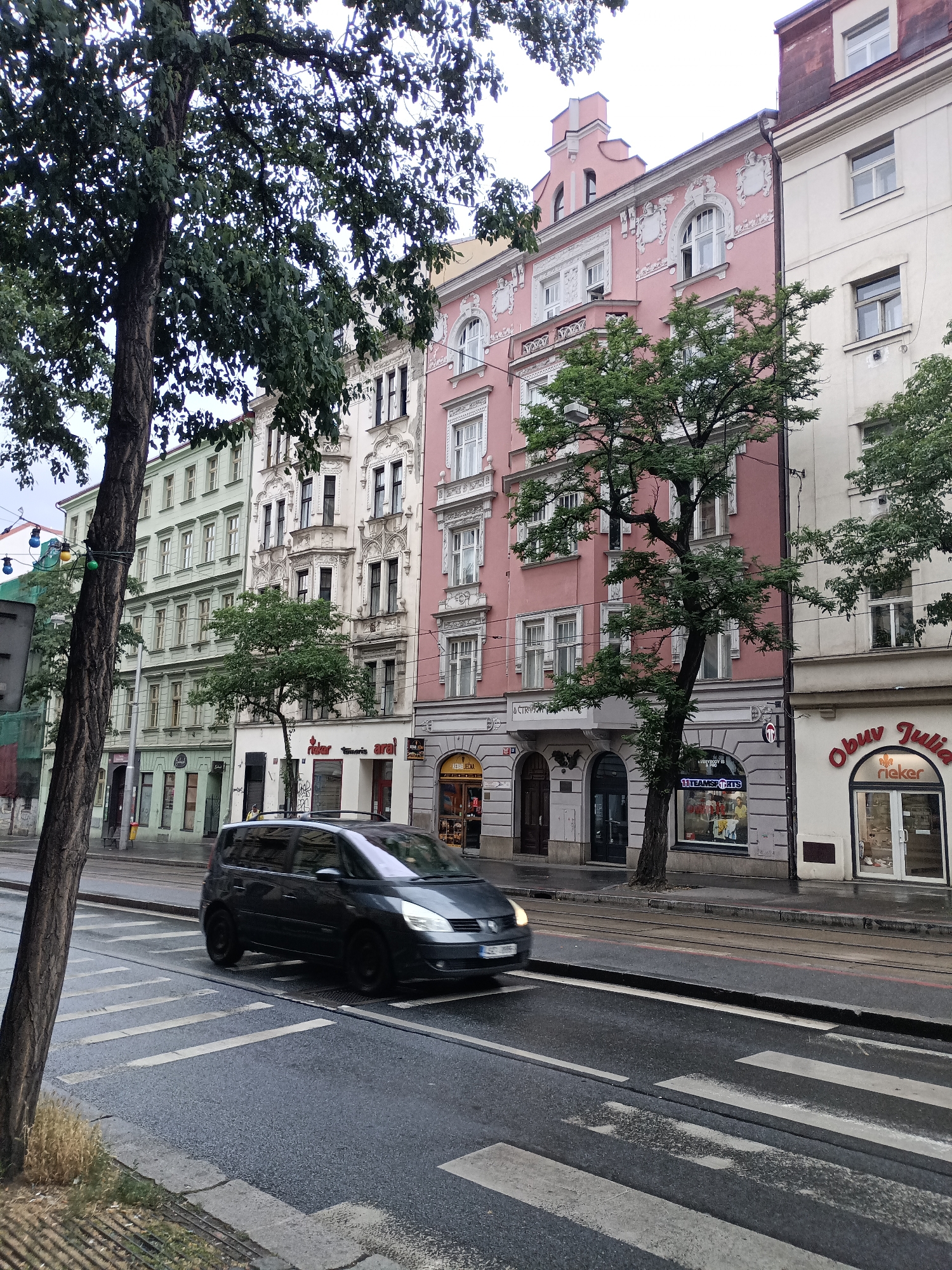 The image captures a street scene in Prague, Czechia, under an overcast sky. In the foreground, a dark-colored minivan is driving across a wet pedestrian crossing. The road appears damp, indicating recent rain or the general humidity of the day. On the right side of the street, a row of buildings lines the sidewalk. These buildings exhibit a mix of architectural styles, with ornate detailing and varied facades. One prominent building is painted pink, featuring arched windows and decorative elements. Another building to its right is a lighter cream color. Several trees with lush green foliage are visible, partially obscuring the buildings. Storefronts are present at street level, with signage such as "Obuv Julia" and "rieker" visible, suggesting retail businesses. The overall atmosphere is that of a typical urban environment.
