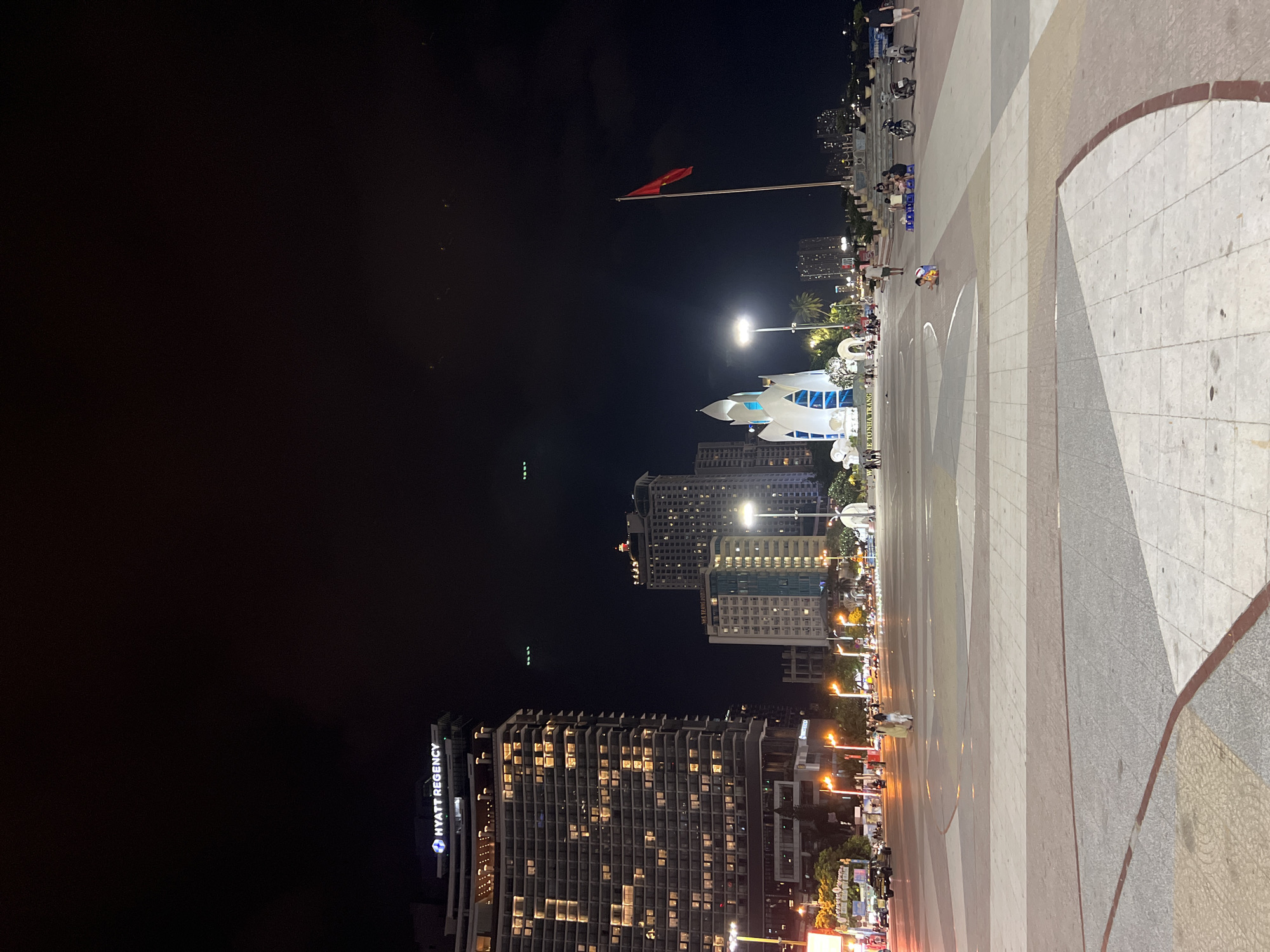 The image depicts a nighttime scene in Nha Trang Ward, Vietnam. In the foreground, there's a paved area with different shades and patterns. In the distance, there are illuminated buildings, including the Hyatt Regency. A Vietnamese flag flies atop a pole. An illuminated swan-like structure can be seen. There are people walking around and vehicles moving along the roads near the buildings. The sky is dark.