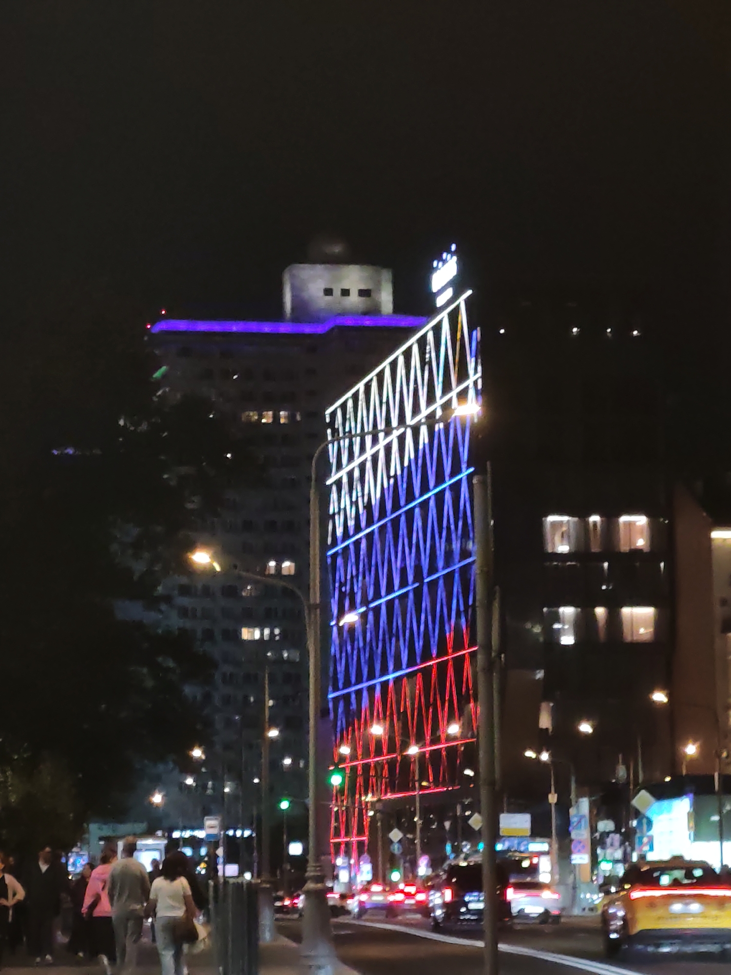 The image depicts a nighttime street scene in an urban environment. A tall building with purple lighting along its upper edge dominates the background, topped by a structure with dark rectangular windows. In the foreground, a large illuminated display panel with a crisscross pattern of red and blue lights stands prominently. The panel is supported by a pole, and a streetlamp with a bright orange light is visible to its left.
To the left of the display, a dark, leafy tree obscures part of a building. A sidewalk runs along the bottom of the image, where several people are walking. One person is wearing a light-colored top and dark pants, another a pink jacket, and a third a white top and grey pants. Further down the sidewalk, more figures are visible.
The street in front of the sidewalk is populated by vehicles. A yellow taxi is the most prominent vehicle on the right side of the road, with its taillights illuminated. Other vehicles, including cars and possibly a bus, are visible in the background, with their headlights and taillights creating streaks of light. Traffic lights, including a green one, are also present. The overall scene suggests a busy city street at night, illuminated by artificial lights from buildings, signs, and vehicles.