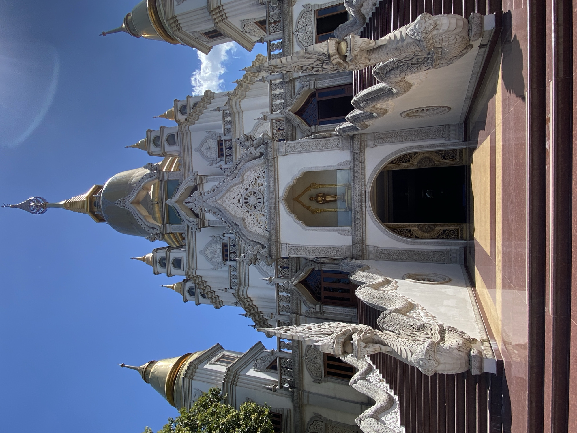 The image captures a low-angle view of a white and gold ornate Buddhist temple structure with intricate detailing. The temple features multiple spires, decorative moldings, and carvings. Two large stone dragons flank the central staircase leading up to the temple entrance. The sky is clear blue with some wispy clouds visible. The building appears to be located in Dĩ An, Vietnam.
