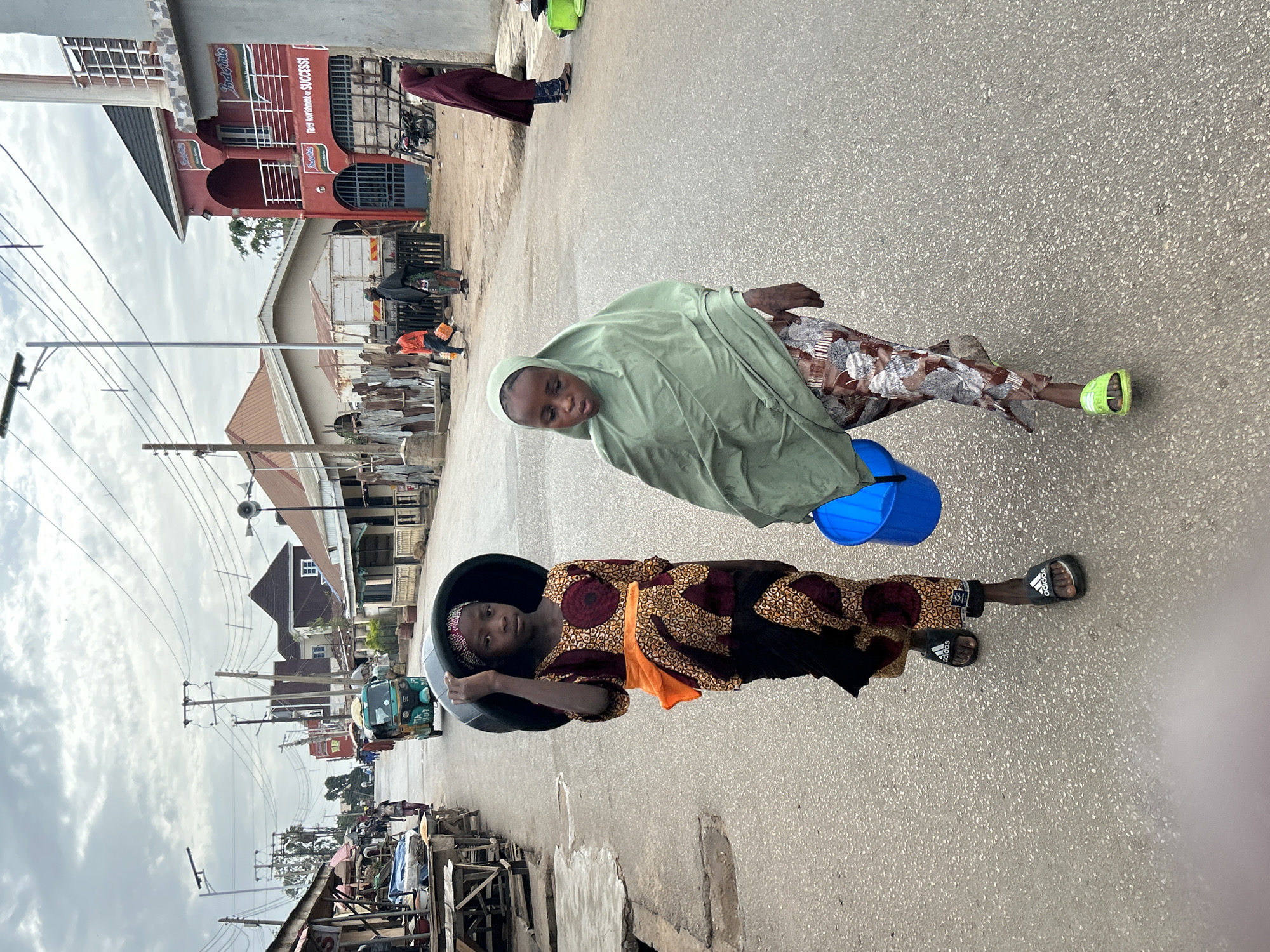 Two young people, likely children, are on a street in Jos, Nigeria. One wears a black-patterned dress with orange accents and carries a black bucket. The other wears a light green headscarf and carries a blue bucket. Buildings and power lines are visible along the street. There are pedestrians and vehicles in the background.