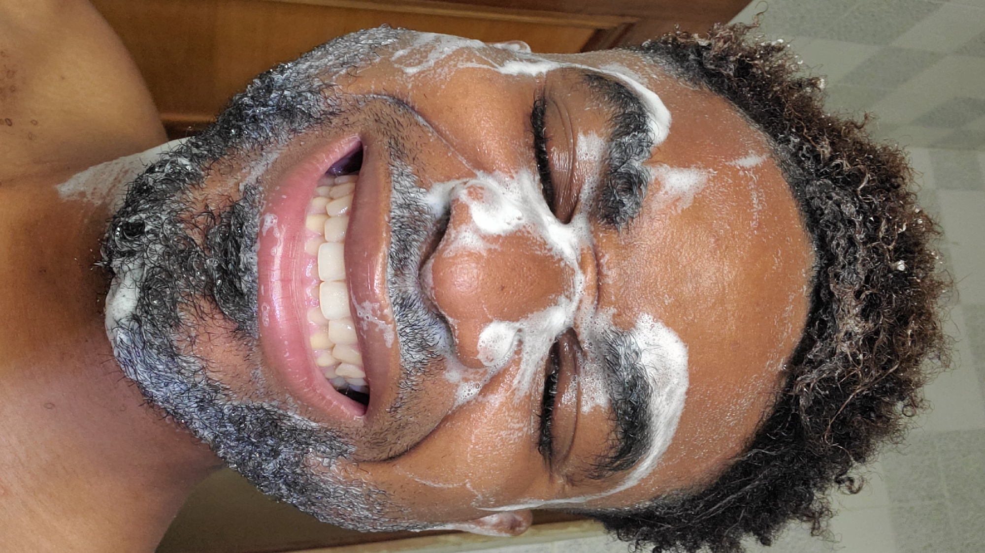 An close-up, rotated portrait shows an individual with dark skin, likely male, immersed in a facial cleansing routine. The person's face, beard, and parts of their curly dark hair are covered in a thick layer of white, foamy lather.
The subject is captured in a moment of apparent joy, displaying a broad, open-mouthed smile that reveals a full set of bright white teeth and pink gums. Their eyes are closed or squinted due to the foam, further emphasizing the cheerful expression. The lather is distributed across the forehead, around the eyes, over the nose, and throughout the beard, indicating a thorough wash.
The background suggests an indoor setting, possibly a bathroom, with muted light and faint tile patterns visible. The image is taken from a slightly upward angle, with the subject's head tilted to the right. This activity, possibly a shower or facial wash, conveys a sense of personal care and a lighthearted, happy demeanor. No discernible text is visible in the image. Given the provided context, this scene takes place in Obosi, Nigeria.