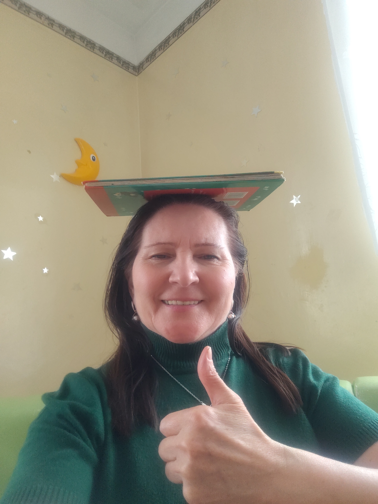 A middle-aged woman poses for a selfie indoors. She is wearing a green turtleneck sweater and has a book balanced on her head. She is smiling and giving a thumbs up. The background shows a yellow wall with star-shaped stickers and a yellow crescent moon wall decoration. The location is seemingly a room in a house, presumably in Sarapul, Russia.