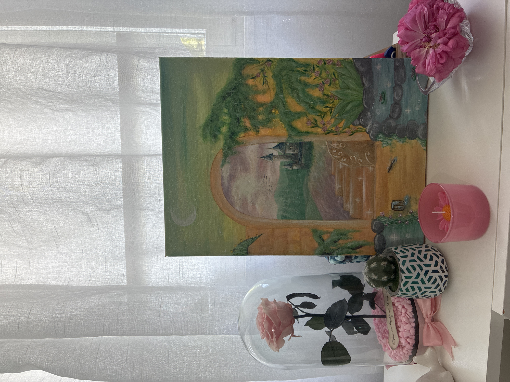 The image shows an indoor scene with a white, sheer curtain in the background. In front of the curtain is a canvas painting depicting a scene with an archway leading to a castle. The painting has green foliage at the top, and a moon is visible on the left side. To the right of the painting, there is a pink flower in a glass vase, a pink candle, and a cactus in a patterned pot. Below the painting, a preserved rose is displayed in a glass dome, sitting atop a base decorated with pink pebbles and a pink ribbon. A wooden spoon is inserted in the pebbles. The scene appears to be set on a white surface, possibly a table or shelf.