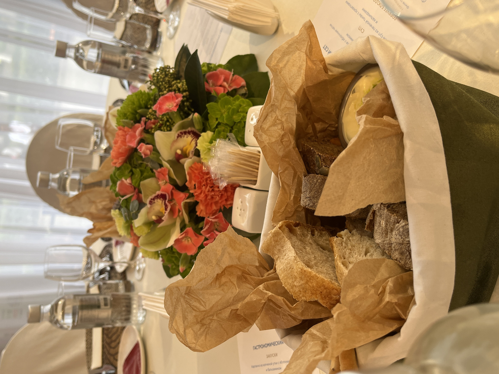 This image captures a well-prepared table setting for a meal or event in Shushary, Russia. The scene is indoors, likely a restaurant or catered venue.
In the foreground, a basket or container lined with green fabric and crumpled brown paper holds an assortment of sliced bread, including light and dark varieties, along with a small cup of butter or dip. Adjacent to this, a vibrant floral centerpiece dominates, featuring orchids, green hydrangeas, and other pink and orange flowers.
The table is neatly set with multiple empty wine glasses, clear water bottles, and partially visible white plates, suggesting place settings for several guests. Small white shakers, likely for salt and pepper, and a stack of wooden disposable cutlery are also present near the floral arrangement.
Several white paper menus are visible on the table. Readable text from the menus includes "ГАСТРОНОМИЧЕСКИЙ" (Gastronomic), "ЗАКУСКИ" (Appetizers), "Суп" (Soup), and "ГОРЯЧЕЕ БЛЮДО" (Hot Dish). Specific menu items mentioned are "нем-суп из печеной тыквы с креветками и кальмарами" (cream soup from baked pumpkin with shrimp and squid) and appetizer components referencing "яблочных бальданинси" (apple balsamic).
No people are visible, suggesting the event is either awaiting guests or currently in progress with guests out of frame. The overall impression is one of a meticulously organized and inviting dining experience.