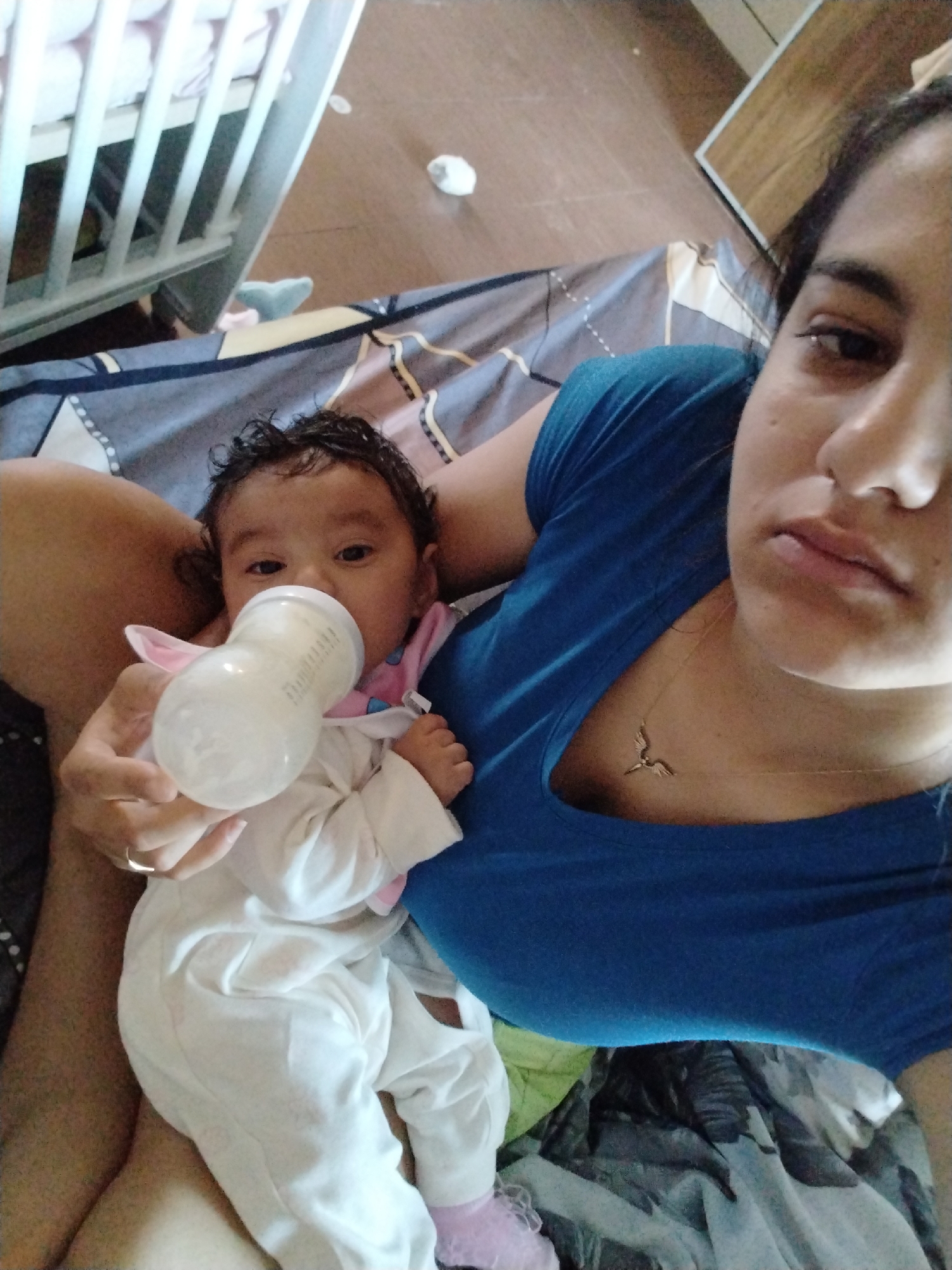 A medium-shot, eye-level photograph depicts a woman holding a baby who is drinking from a bottle. The location is an indoor space, possibly a home, city of Unknown, Unknown. The woman is fair-skinned with dark hair pulled back. She is wearing a blue shirt and a necklace. She looks towards the camera with a neutral expression. In her left hand, she holds a baby, approximately 6-12 months old, who is drinking from a bottle. The baby is wearing a white onesie with pink socks. To the left of the woman and baby, there is a white crib. The background consists of bedding in neutral and grey tones. A piece of furniture, likely a dresser or cabinet, is visible in the background on the upper right. A small, crumpled white object is located on the floor near the crib.