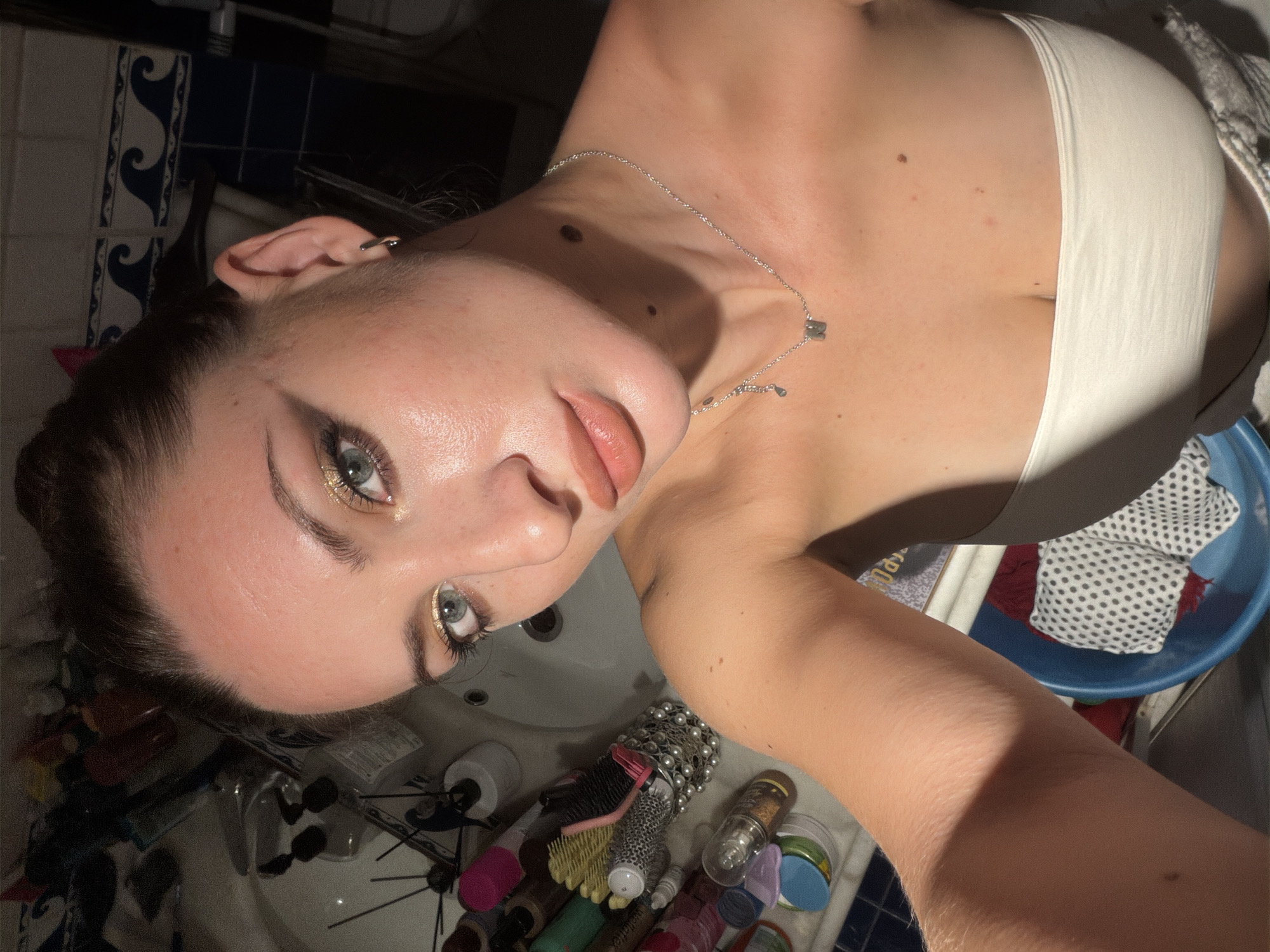 A young woman with dark brown hair pulled back is taking a selfie in a bathroom. She is wearing a white strapless top and a silver necklace with a small charm. She has on makeup, including gold eyeshadow and lipstick. Various toiletries are visible in the background on a bathroom counter. The environment suggests a domestic setting, potentially in Sharm El Sheikh, Egypt, given the blue and white wave pattern tiles on the wall. The lighting is dim and appears to be from artificial sources, casting shadows.
