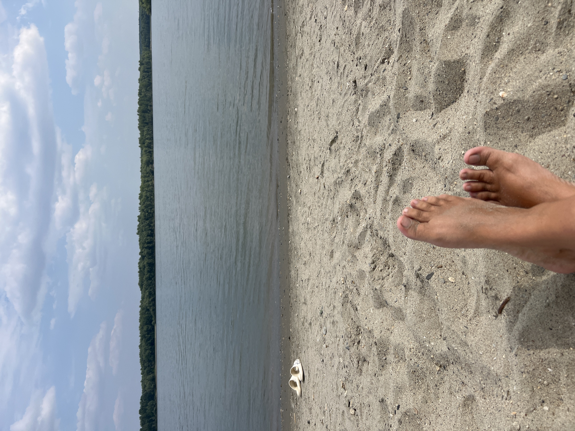 The image depicts a tranquil beach scene on a sunny day. In the foreground, a pair of bare feet are propped up on the sandy shore, with white slide sandals resting nearby. The sand is a light tan color and appears to be composed of fine grains mixed with small pebbles. The water of a lake or river is visible in the mid-ground, with gentle ripples on its surface. Beyond the water, a dense line of green trees forms a natural backdrop, separating the water from a partially cloudy blue sky. The location is identified as Mochishche, Russia, a settlement within the Moscow Oblast known for its natural beauty, including its proximity to the Oka River, which may be the body of water shown in the image.