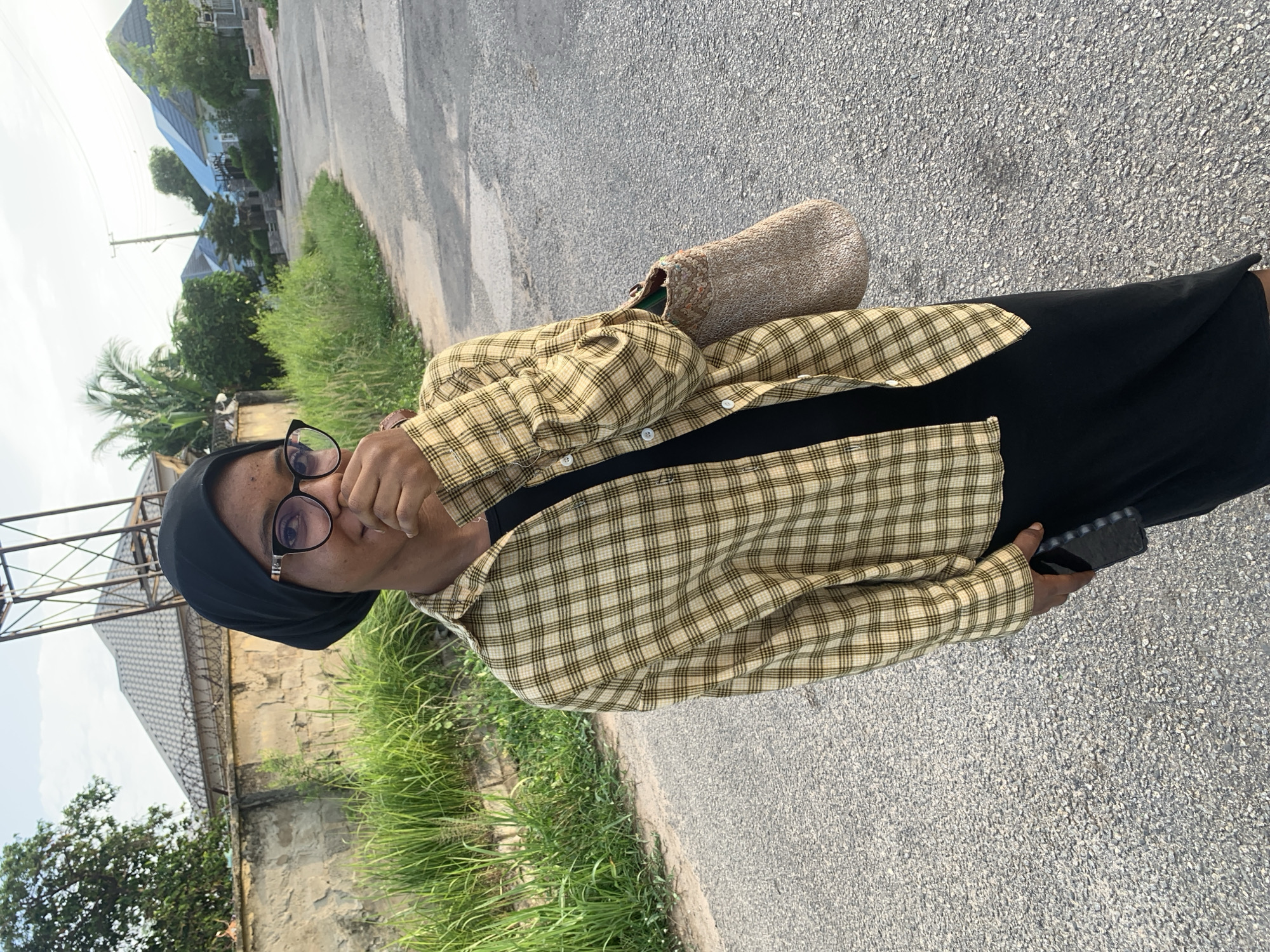 A woman wearing a black headscarf and glasses, a black dress, and a yellow and tan plaid shirt stands on an asphalt road in an urban area. She has her hand to her face, and is holding a black mobile phone in her other hand. She has a woven purse or bag on her shoulder. To her left is a grassy area, a low wall, and a building with a tall metal structure nearby. In the background, are other buildings with blue roofs. The location is presumed to be Abuja, Nigeria, based on the prompt.