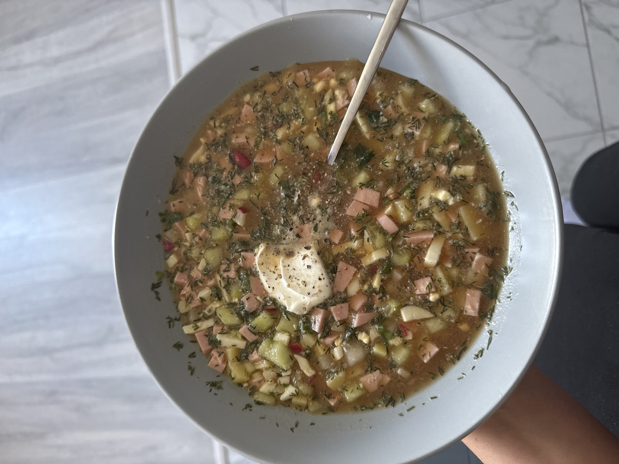 A top-down, close-up photograph displays a gray bowl filled with a cold, savory soup, likely Okroshka, against a light-colored, tiled background. A metal spoon rests in the soup.
The soup features a light brown or tan broth and is abundant with diced ingredients. Visible components include numerous small, pinkish-beige cubes of what appears to be ham or cooked sausage, and diced light green cucumber. Other diced ingredients, possibly potato or hard-boiled egg, are also present. The soup is generously garnished with finely chopped green herbs, predominantly dill, giving it a vibrant texture.
A prominent dollop of a white, creamy topping, likely sour cream or mayonnaise, sits near the center of the bowl, lightly dusted with black pepper. A person's arm or leg, clothed in dark fabric, is partially visible in the bottom right corner, suggesting the bowl is being held or resting on a lap. The background consists of light gray and white tiles with subtle veining, possibly mimicking marble. There is no visible text in the image. The scene suggests a meal prepared and ready to be consumed.