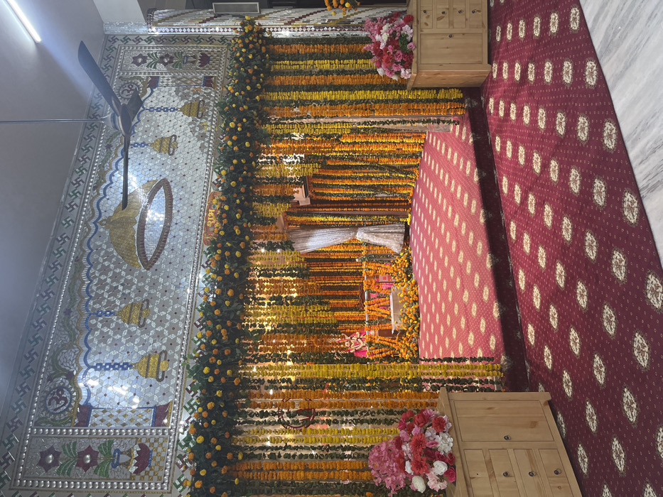 The image depicts the entrance to a temple, located in Panchkula, India, at coordinates 30.723, 76.862. The entrance is adorned with garlands of yellow flowers and green leaves, creating a decorative archway.  Behind the garlands, a large, intricately designed mosaic wall can be seen. The wall features a central decorative motif, with a white ceiling fan above.  A burgundy-colored carpet covers the floor, leading to a small raised platform adorned with more garlands. A single figure is visible, sitting within the inner sanctum of the temple.  Two wooden cabinets with drawers stand on the right side of the image, each containing flower arrangements.  The scene is brightly lit and captures a sense of spiritual devotion.