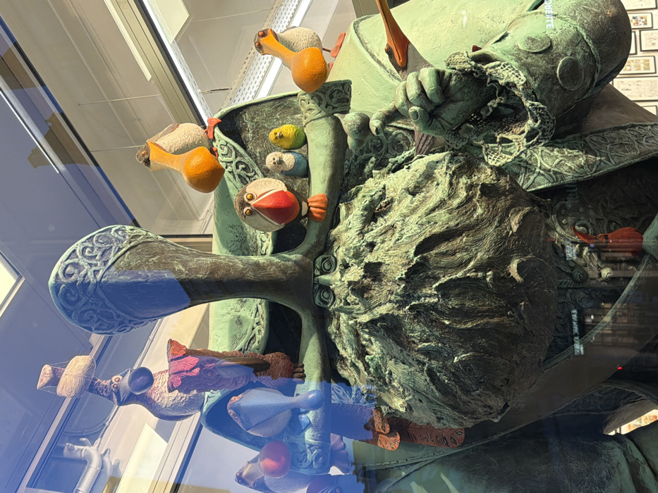 A whimsical bronze sculpture presents a figure with bird-like characters incorporated within its structure. Birds resembling puffins and other species adorn the head, shoulders, and body. The figure, possibly a knight or historical persona, is depicted in armor, with a lace-like detail at the wrist. The sculpture's patina exhibits shades of green and brown, adding to its aged and artistic appearance. The base shows reflections of objects behind the sculpture, providing context.