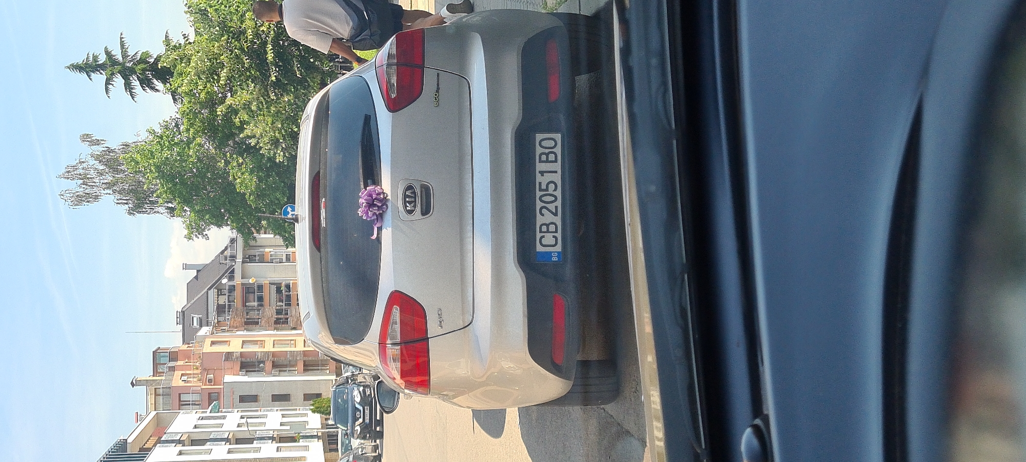 The image shows the rear of a silver Kia Rio parked on a street in Sofia, Bulgaria. A purple bow is affixed to the rear window. The license plate reads "CB 2051 BO". Buildings and trees are visible in the background. A person is walking away from the camera on the right side of the image. The scene appears to be a sunny day.