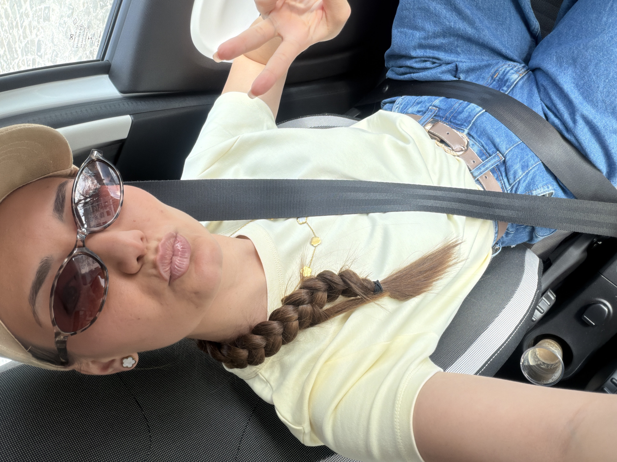 A female individual is depicted from the upper torso, seated in the passenger seat of a vehicle, taking a selfie. She wears a beige baseball cap, brown-tinted tortoise-shell framed sunglasses reflecting trees and buildings, a light yellow short-sleeved t-shirt, blue jeans, and a light brown belt with a metallic buckle. Her dark brown hair is styled in a single braid extending over her left shoulder. White flower-shaped stud earrings are visible, as is a delicate gold-colored necklace with small charms. Her lips are puckered, and her raised right hand displays a peace sign. A black seatbelt is positioned across her chest and lap. The vehicle's interior features dark grey patterned fabric on the seat and black plastic trim. A clear plastic cup containing a light brown substance is in a cup holder in the center console. Inverted text on a sticker visible through the car's passenger window includes "АВТОМОТИВ АД" and "SOFIA", confirming the location is within the city of Sofia, Bulgaria.