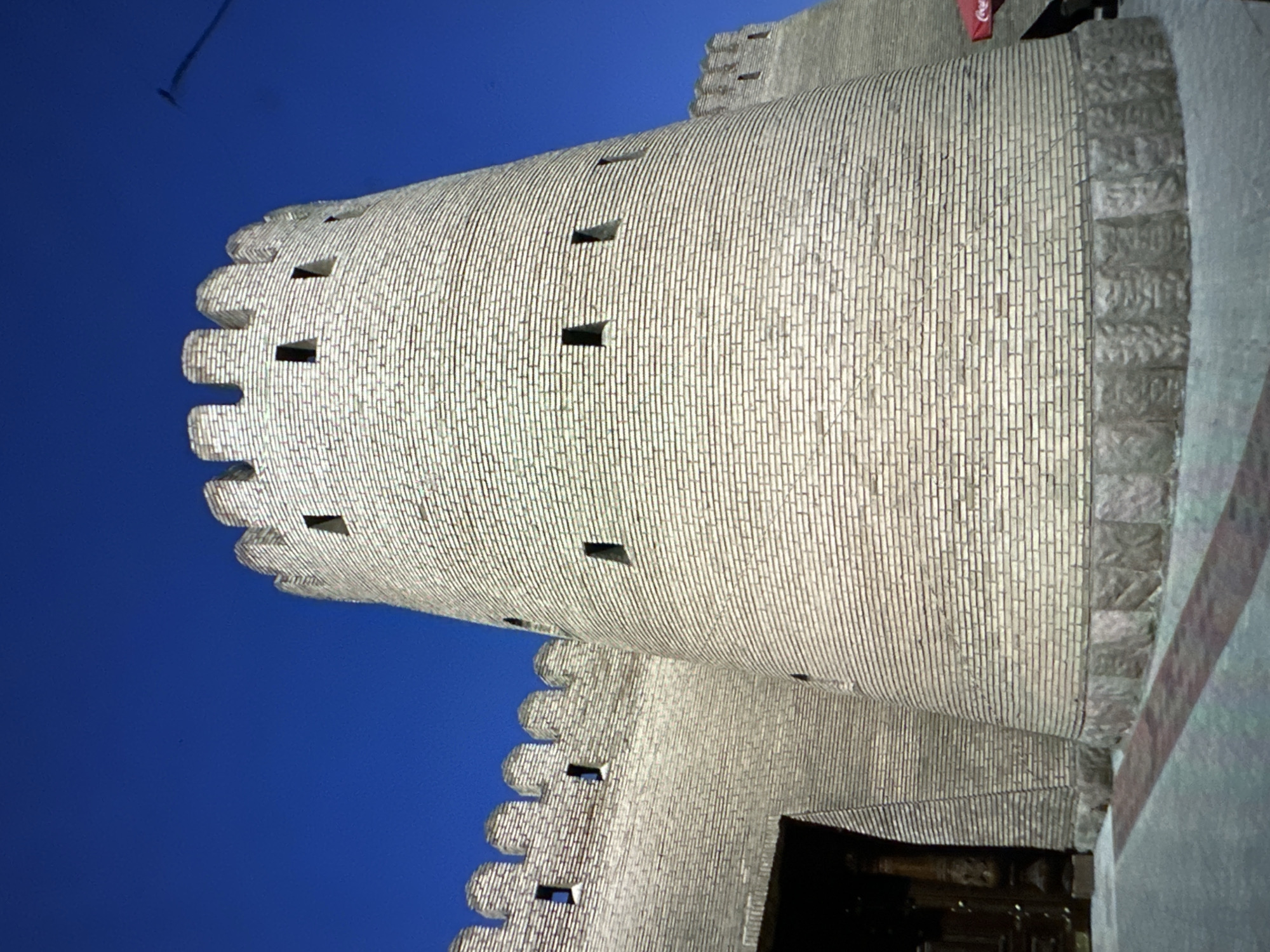 The image shows a close-up, low-angle view of a cylindrical brick tower with crenellations against a clear blue sky. The tower is constructed of light-colored bricks and has several small, rectangular openings. To the left of the tower, a section of a crenellated wall is visible, as well as a dark wooden doorway. The base of the tower is adorned with decorative stone blocks. In the background, another crenellated structure is visible, and a portion of a red Coca-Cola umbrella can be seen to the right. The scene appears to be in an outdoor location, possibly a historical site or fortress in the city of Кызыл-Булак, Kyrgyzstan.