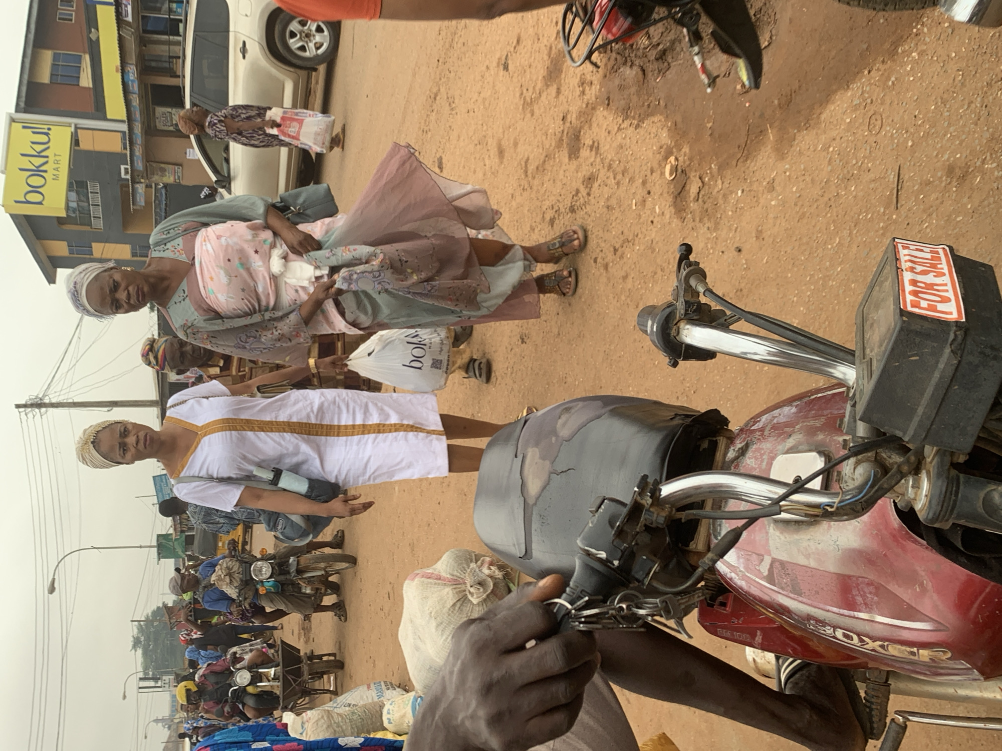 In the city of Shagamu, Nigeria, a bustling street scene unfolds. The foreground is dominated by the front of a red motorcycle, its handlebars and part of the fuel tank visible. A black box attached to the motorcycle displays a red and white "FOR SALE" sticker. A person's dark-skinned hand, holding keys, rests on the handlebars. In the middle ground, two women walk across the dirt road. The woman on the left wears a white dress with a gold trim and carries a light blue bag. The woman on the right is dressed in a flowing, light-colored patterned outfit and holds a light blue bag and a fabric. Behind them, several individuals are present. One man, wearing a yellow helmet, rides a motorcycle that is towing a cart. Another man on a motorcycle is also visible. A gold-colored SUV is parked on the right side of the road. In the background, a building with a yellow sign reading "bokku! MART" is prominent. The environment appears to be a marketplace, with goods in sacks visible on the left side of the image. The ground is covered in dry, reddish-brown dirt. The sky is overcast, suggesting cloudy weather.