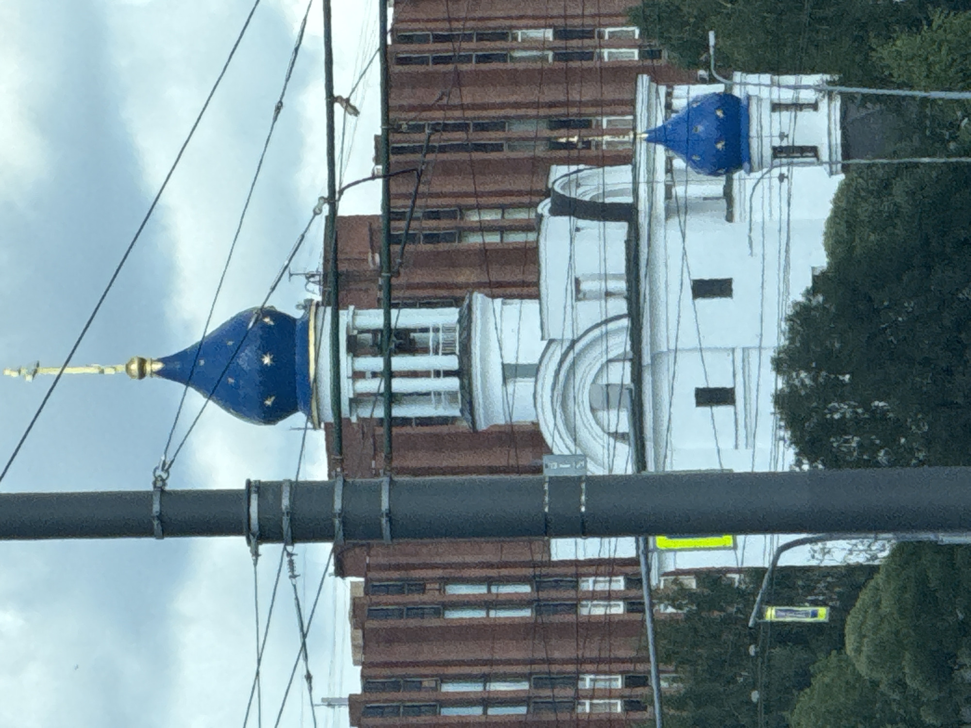 This image shows a view of a white building with a blue domed roof topped with a golden cross. The building appears to be an Orthodox church, with architectural details such as arched windows and columns. Behind the church, a tall, brick building with many windows is visible. The scene is framed by power lines and a large, dark gray utility pole in the foreground. Trees and a traffic light are also present, suggesting an urban or semi-urban environment. The sky is overcast with a grayish-blue hue. The location is identified as Sinyavino, Russia. There are no people visible in the image.