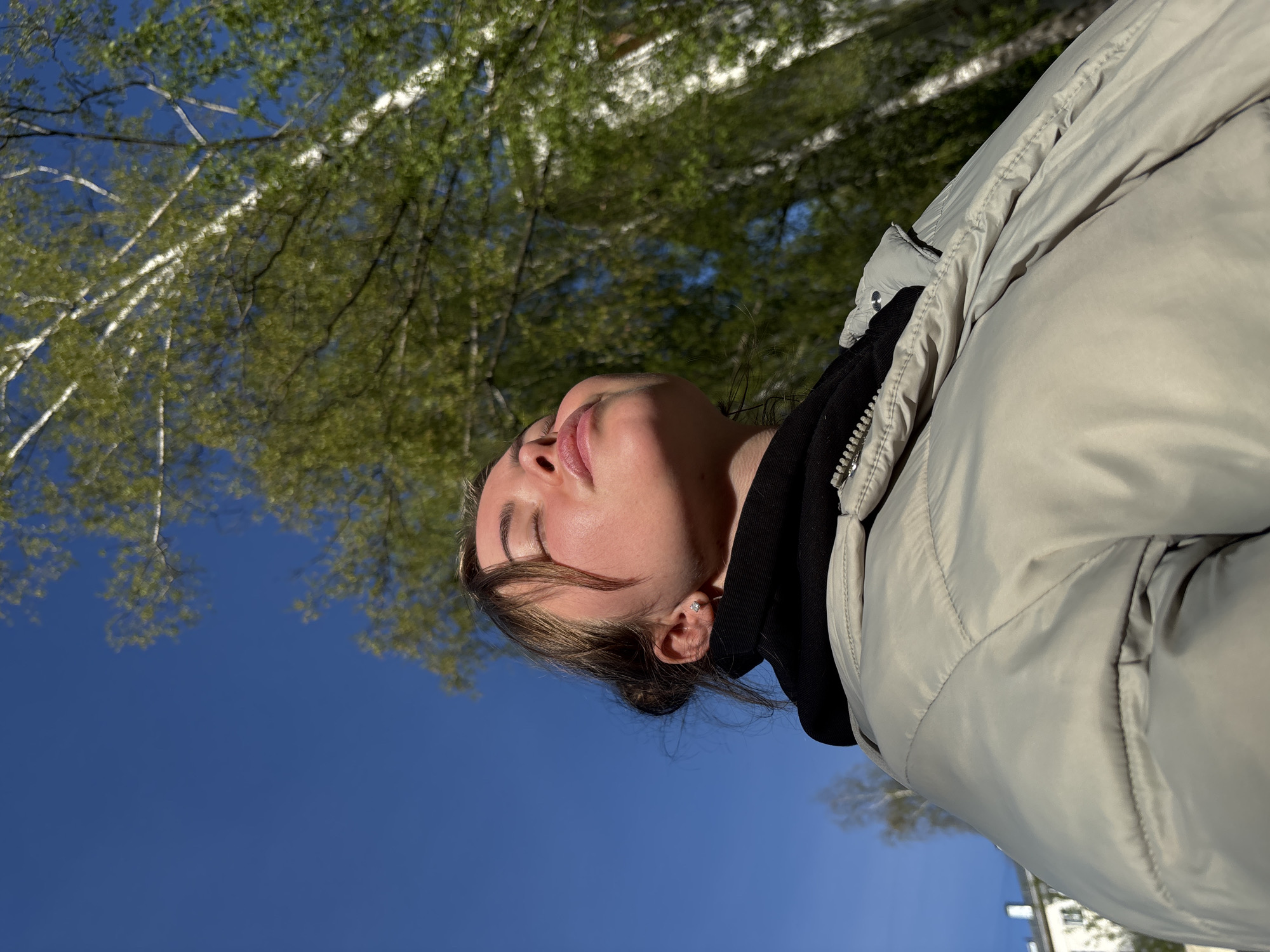 A young woman, seen from a low angle, faces upwards toward the sky. She is wearing a light-colored puffer jacket with a black hooded sweatshirt underneath. Her eyes are closed, and her lips are slightly parted. Her hair is pulled back. Above her are the branches and leaves of trees against a bright blue sky. At the very bottom of the image, hints of a building are visible. The location is determined to be Izhevsk, Russia based on the tree type (Birch) and a building type (low level urban apartment) common to the Izhevsk, Russia region.