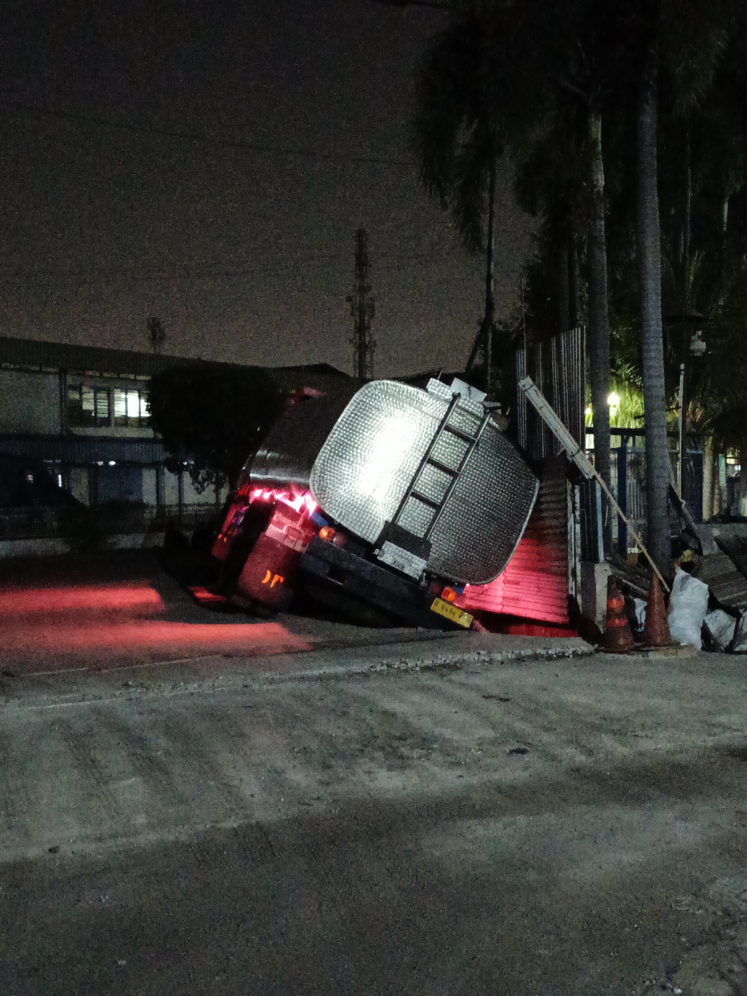 This image shows a tanker truck that has overturned at night, located in the city of East Jakarta, Indonesia. The tanker, which appears to be made of metal with a textured surface, is lying on its side, with its rear wheels lifted off the ground. Red lights from the truck's tail lights cast a glow on the road and the surrounding area. A section of a building with illuminated windows is visible in the background, along with some palm trees. Several orange traffic cones and a white bag are placed near the overturned vehicle. The ground is a mix of asphalt and gravel, with tire tracks visible. The scene appears to be illuminated by artificial lights from the surrounding environment.
