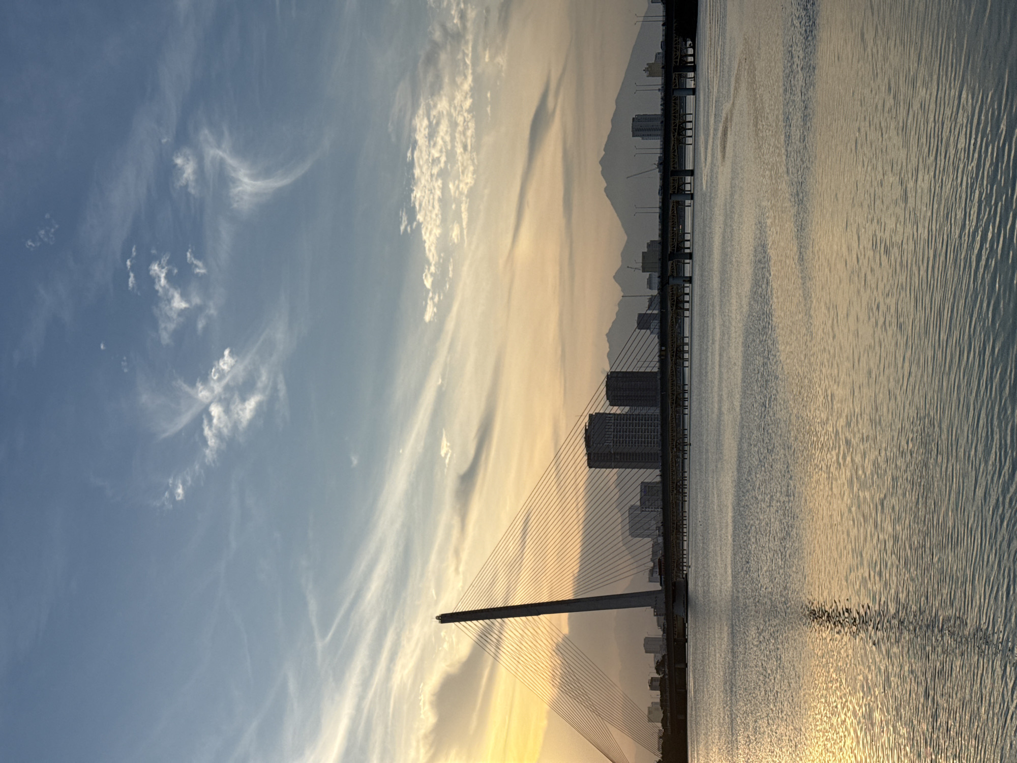 The image depicts the Han River bridge in Da Nang, Vietnam at sunset. A tall, slender pylon of the cable-stayed bridge dominates the left side of the frame, with numerous cables extending to the bridge deck. In the background, a city skyline with several modern buildings is visible, with a range of mountains forming a backdrop beyond the city. The sky above is a gradient of blue and warm sunset colors, with wispy clouds. The Han River occupies the foreground, reflecting the colors of the sky and the bridge. No people or actions are discernible in the image.