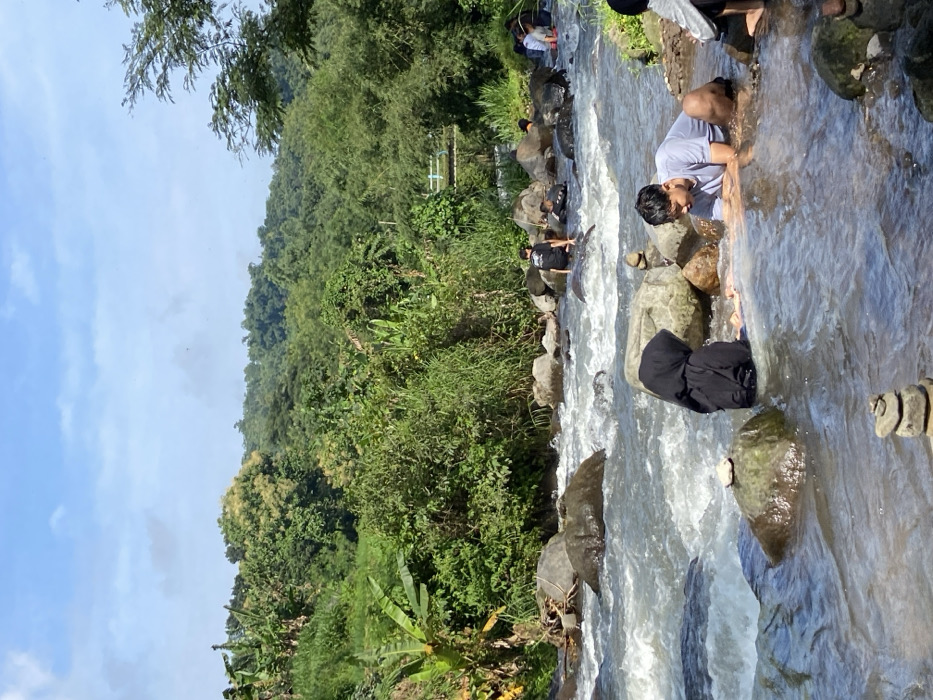 A group of people are in a river, some standing on rocks. A man in a light-colored shirt leans over the water. Another person, wearing a black hooded garment, is partially submerged. Lush green vegetation covers the banks of the river and extends up a hillside. A blue sky with scattered clouds is visible above the trees. The river appears shallow, with visible rocks and rushing water.