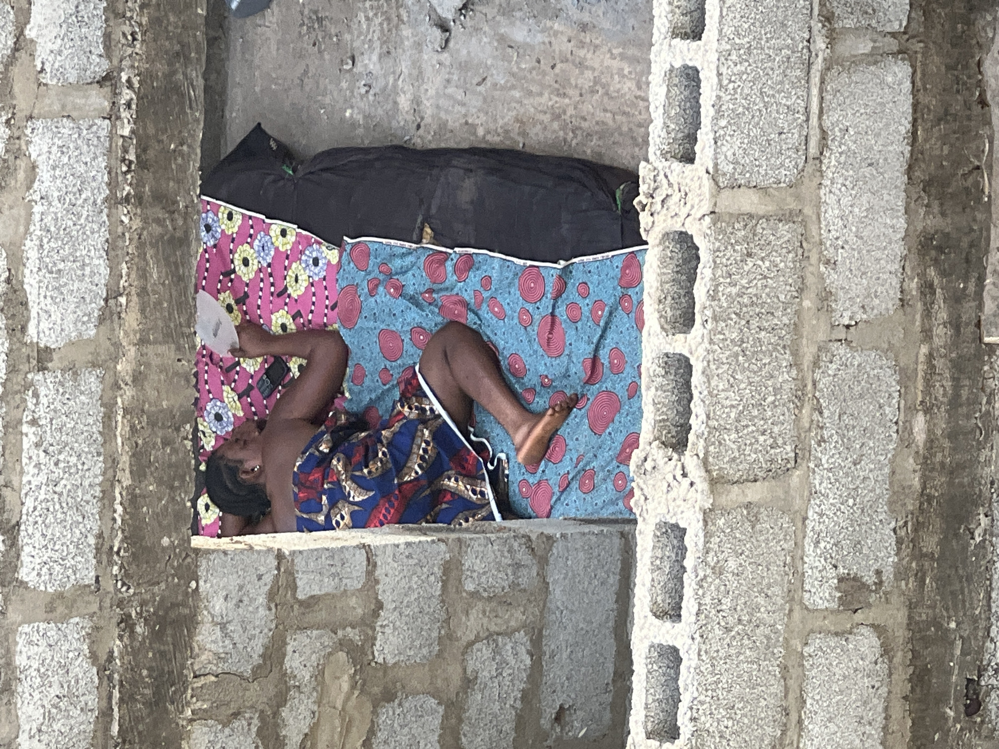 An overhead shot depicts a person lying on a bedroll situated within a concrete structure, likely in Abuja, Nigeria, suggested by the cinderblock construction style. The person is lying on a bedroll with two patterned textiles, one with pink and floral designs and the other with blue and red circular motifs, covered partially by a dark blanket. The individual is covered partially by a patterned cloth. The structure appears to be in progress, as suggested by the exposed cinderblocks.