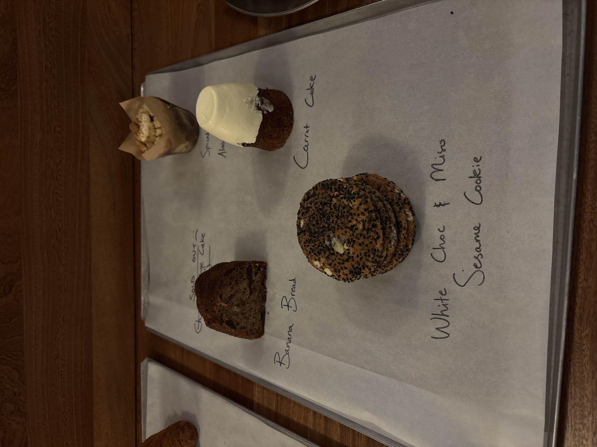The image shows a tray of baked goods, likely in a bakery or cafe, with handwritten labels. On the left side of the tray, there's a small paper bag containing "Spiced Alm" (presumably Spiced Almond), a cake labeled "Carrot Cake" with white frosting, a slice of "Banana Bread," and a stack of "White Choc & Miso Sesame Cookie." A note "SOLD" is written above what appears to be "Orange Cake." The tray rests on a wooden surface. The location details are not discernible from the image.