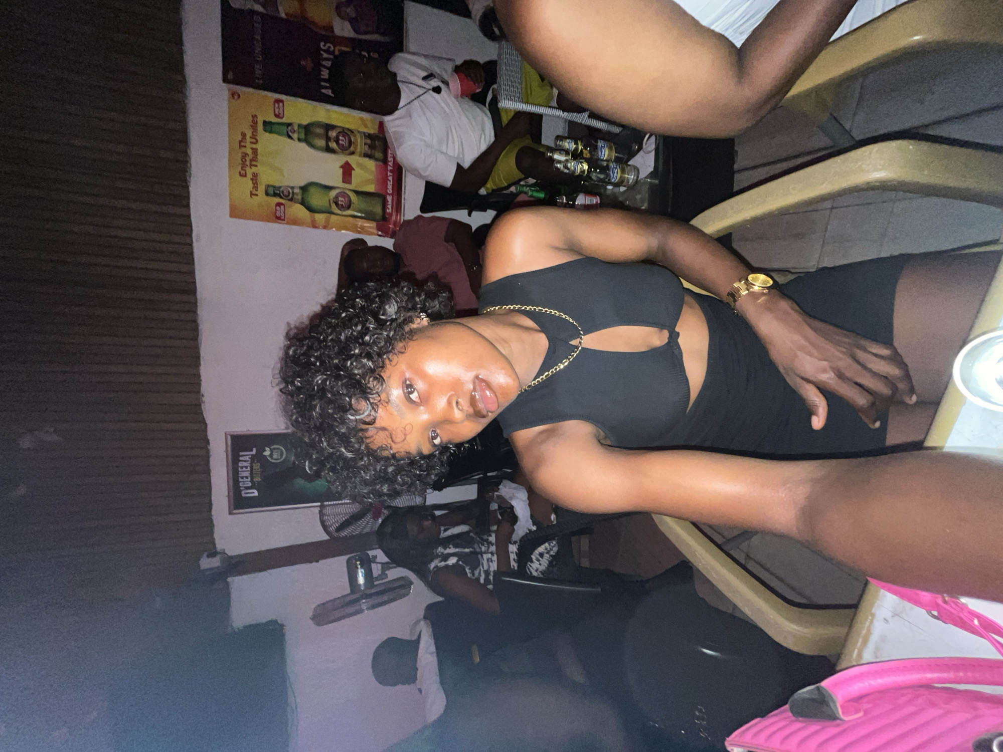A cluttered indoor space in Ado, Nigeria, shows a woman in a black dress with curly hair and a gold watch posing for a selfie. Other patrons are seated at nearby tables with bottles of alcoholic beverages. Posters of "33" Export Beer and "D'General Bitters" are visible on the wall. The lighting is dim, creating shadows and highlighting the reflective surfaces.
