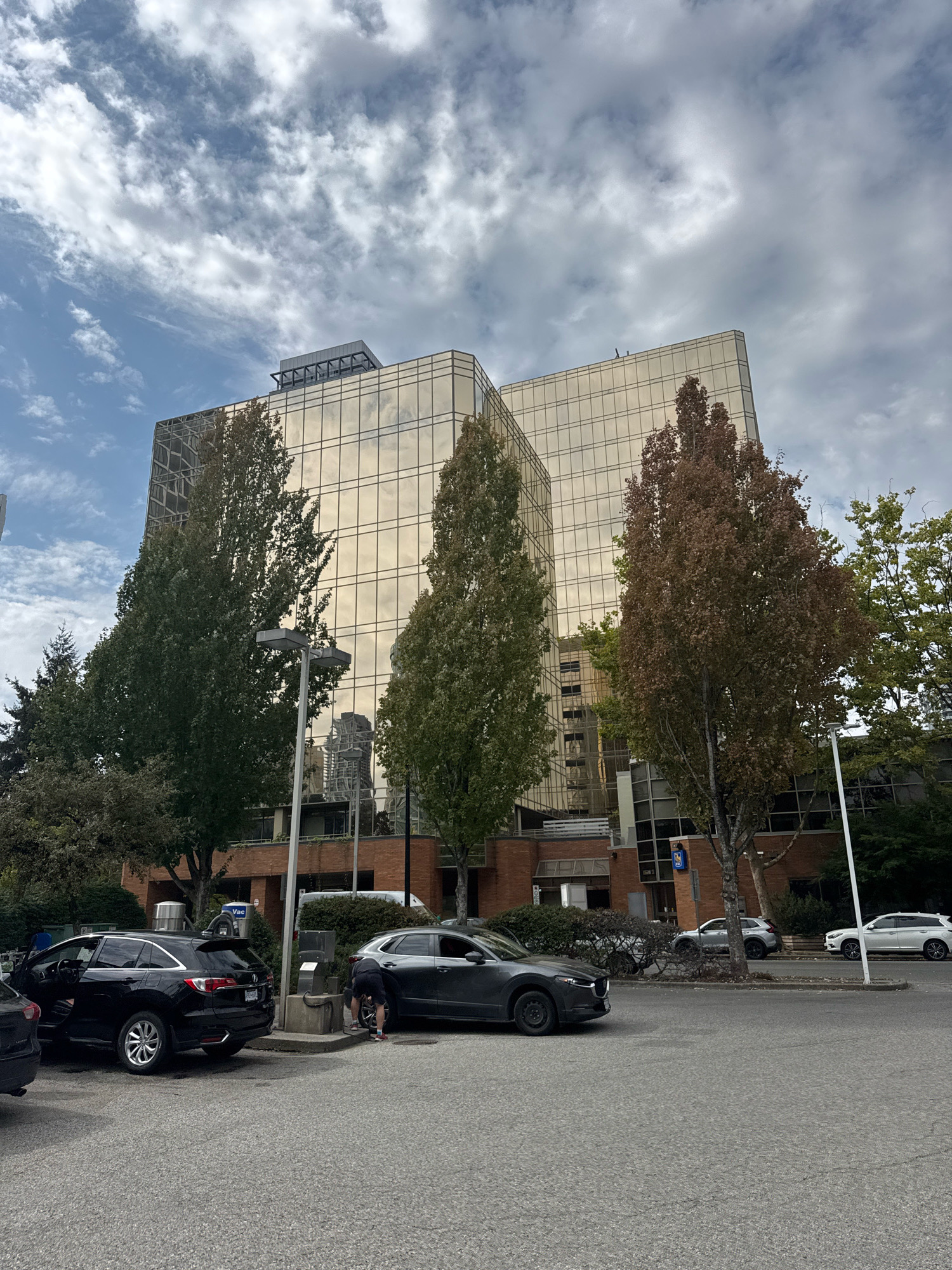 The image depicts a scene in front of a modern glass-covered office building located at 4500 Willingdon Avenue in Burnaby, Canada. The building has a distinctive golden facade that reflects the sky and surrounding environment. In the foreground, a grey sedan is parked next to a black SUV. A person, dressed in dark shorts and a dark t-shirt, is bent down near the rear of the black SUV, appearing to be engaged in cleaning or detailing the vehicle. The parking lot is paved, and a street with several cars parked along it is visible to the right of the building. Trees with green and brown foliage are interspersed around the building and along the street, suggesting it is autumn. The sky is partly cloudy, with a mix of blue sky and white clouds.