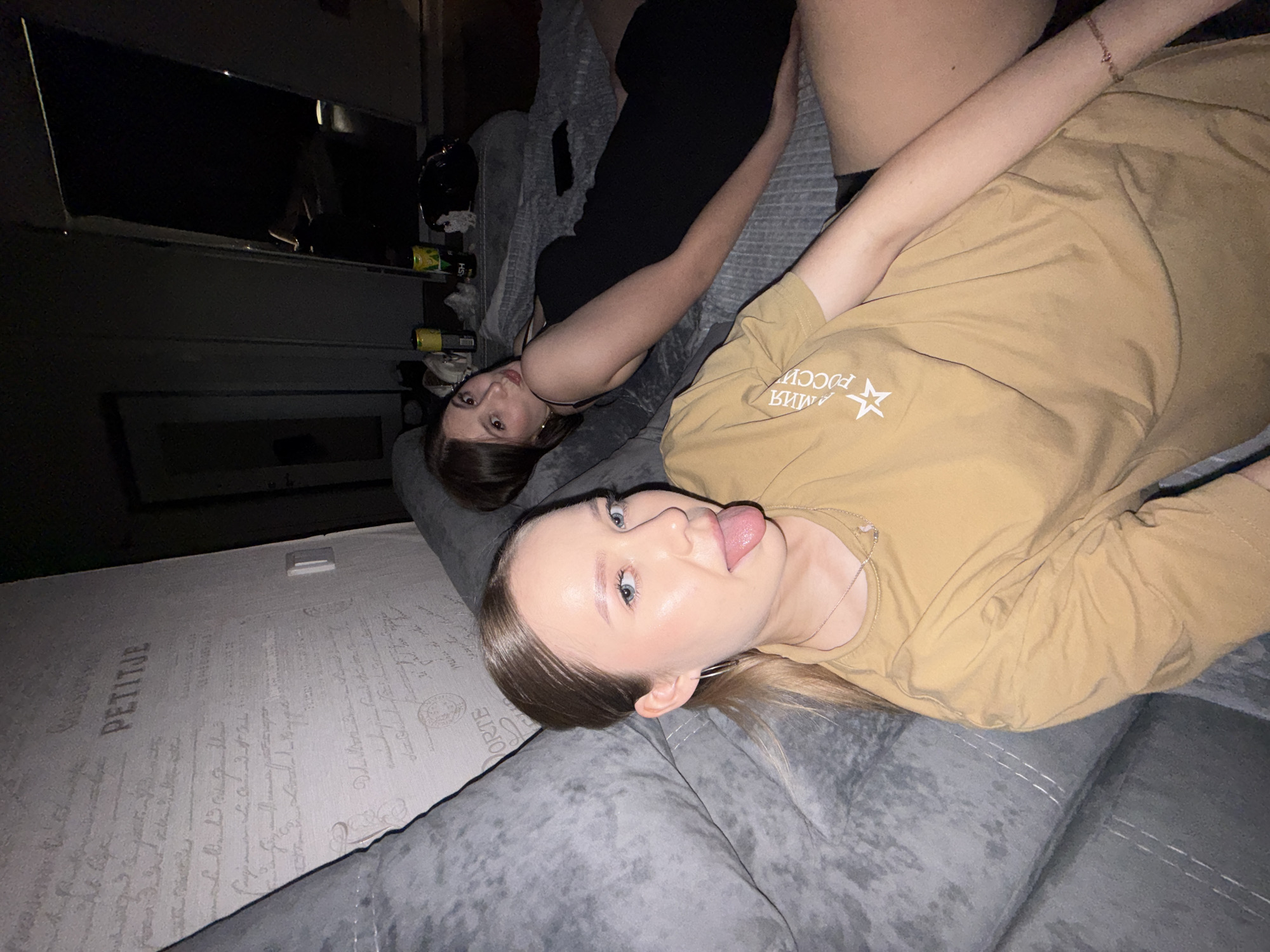 In the image, two young women are lounging on a large, grey sectional sofa in an indoor setting. The woman in the foreground, with blonde hair pulled back in a ponytail, wears a mustard-colored t-shirt and is sticking out her tongue at the camera. She has blue eyes and light skin, and is wearing hoop earrings and a delicate silver necklace. The woman in the background, with dark brown hair, is wearing a black dress and is looking over her shoulder towards the camera. The room appears to be dimly lit, with a wall covered in a textured wallpaper featuring decorative script. A mirror is visible in the background, reflecting a dark space. On a surface behind the sofa, several cans and a black bag are scattered. The overall scene suggests a casual gathering or a moment of relaxation.