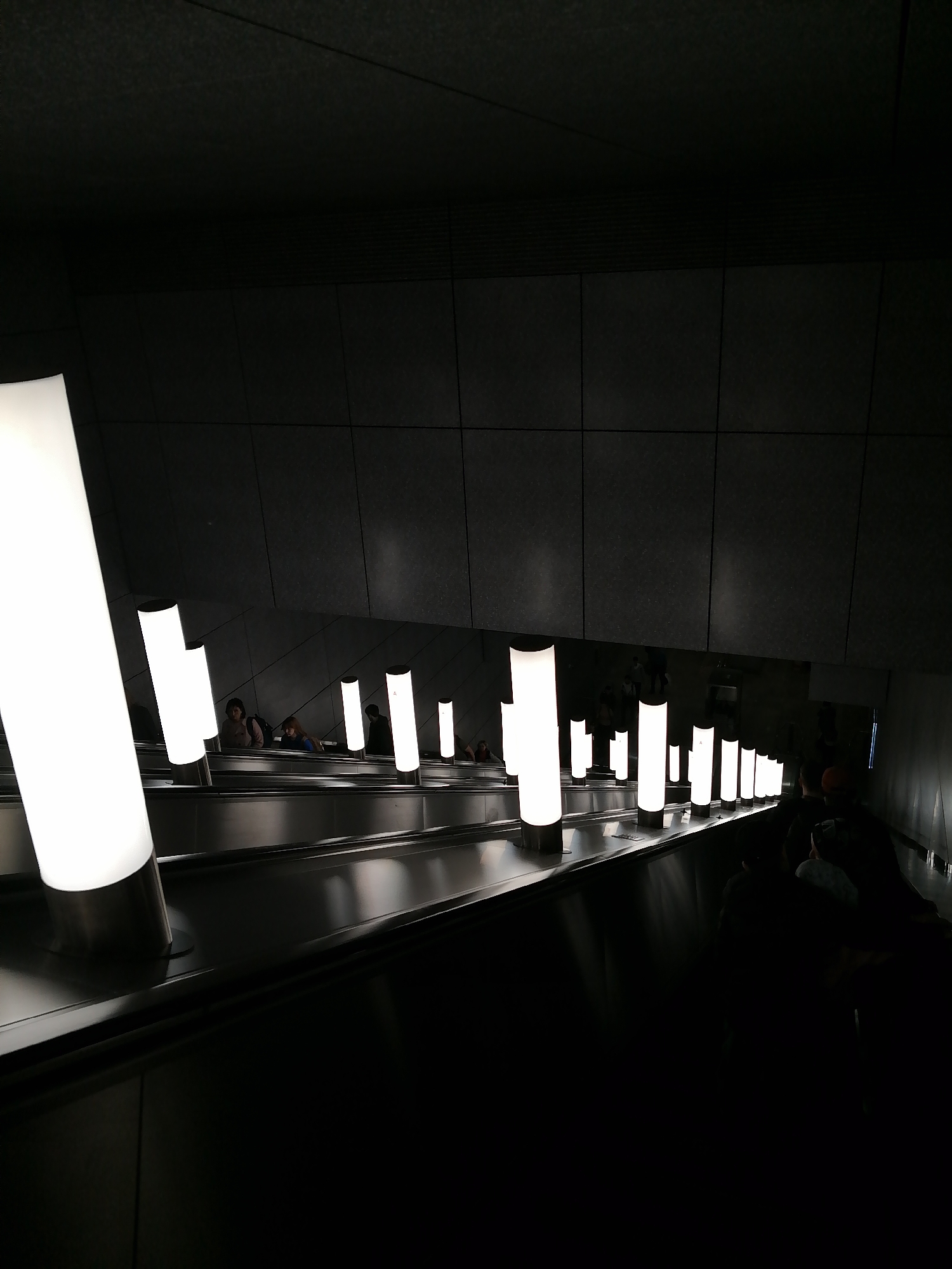 The image depicts a descending escalator in a dimly lit, modern-looking underground space, likely a subway station. People are riding the escalator in both directions. The escalator is lined with evenly spaced, tall, cylindrical lamps that emit a bright, white light, creating a contrast against the dark environment. The walls are composed of dark, rectangular panels. The location is Unknown, Unknown.