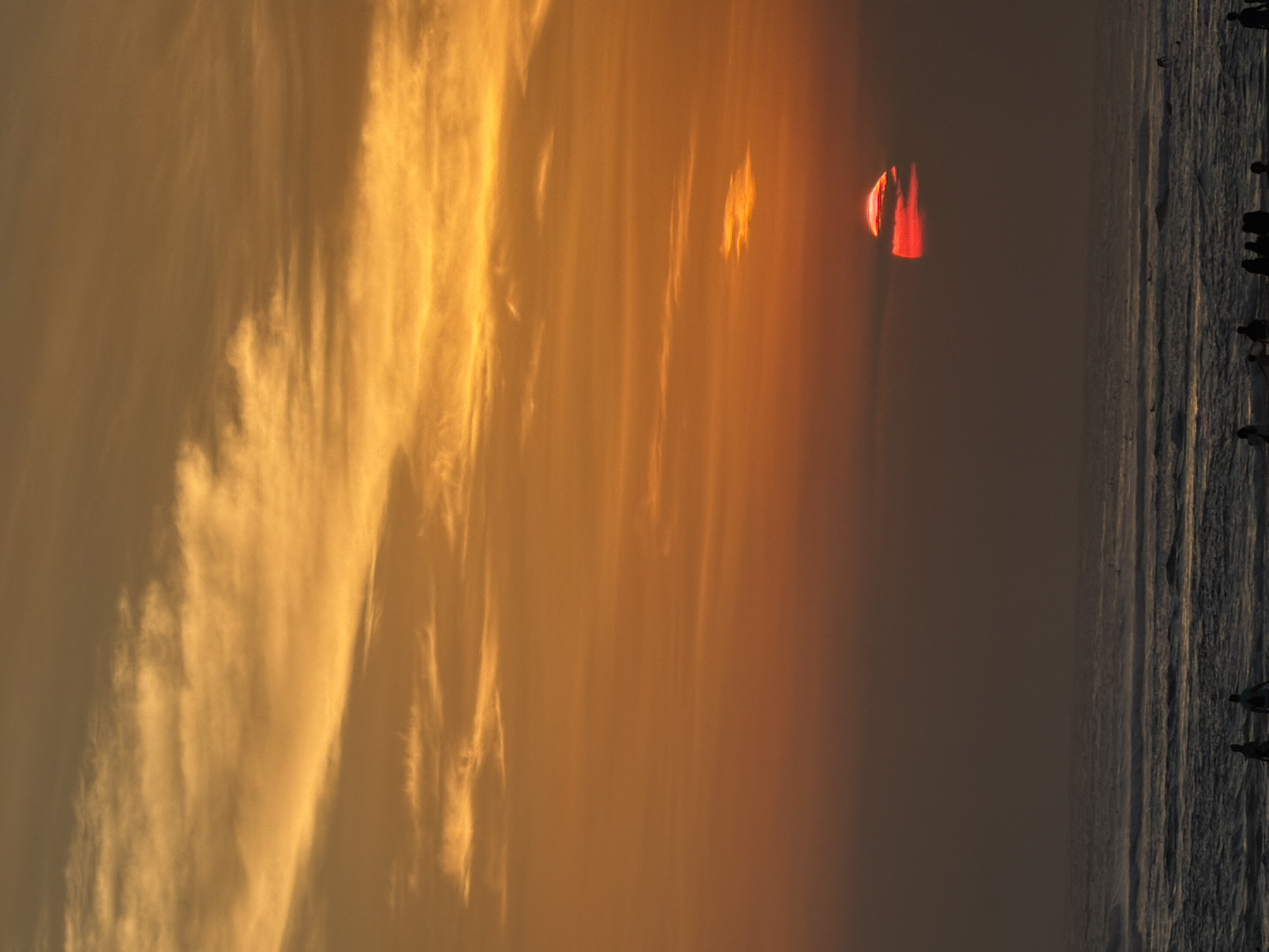 This is a vertical, low-angle shot of the ocean at sunset in North Kuta, Indonesia. The sky is a gradient of orange and yellow, with wispy clouds. The sun, a vibrant red orb with some clouds partially obscuring it, is low on the horizon. The ocean is choppy with white waves. Silhouettes of people are visible on the beach in the foreground. Some individuals appear to be walking, while others are standing. A few surfers are in the water. The overall atmosphere is serene and depicts a typical sunset scene on a beach.