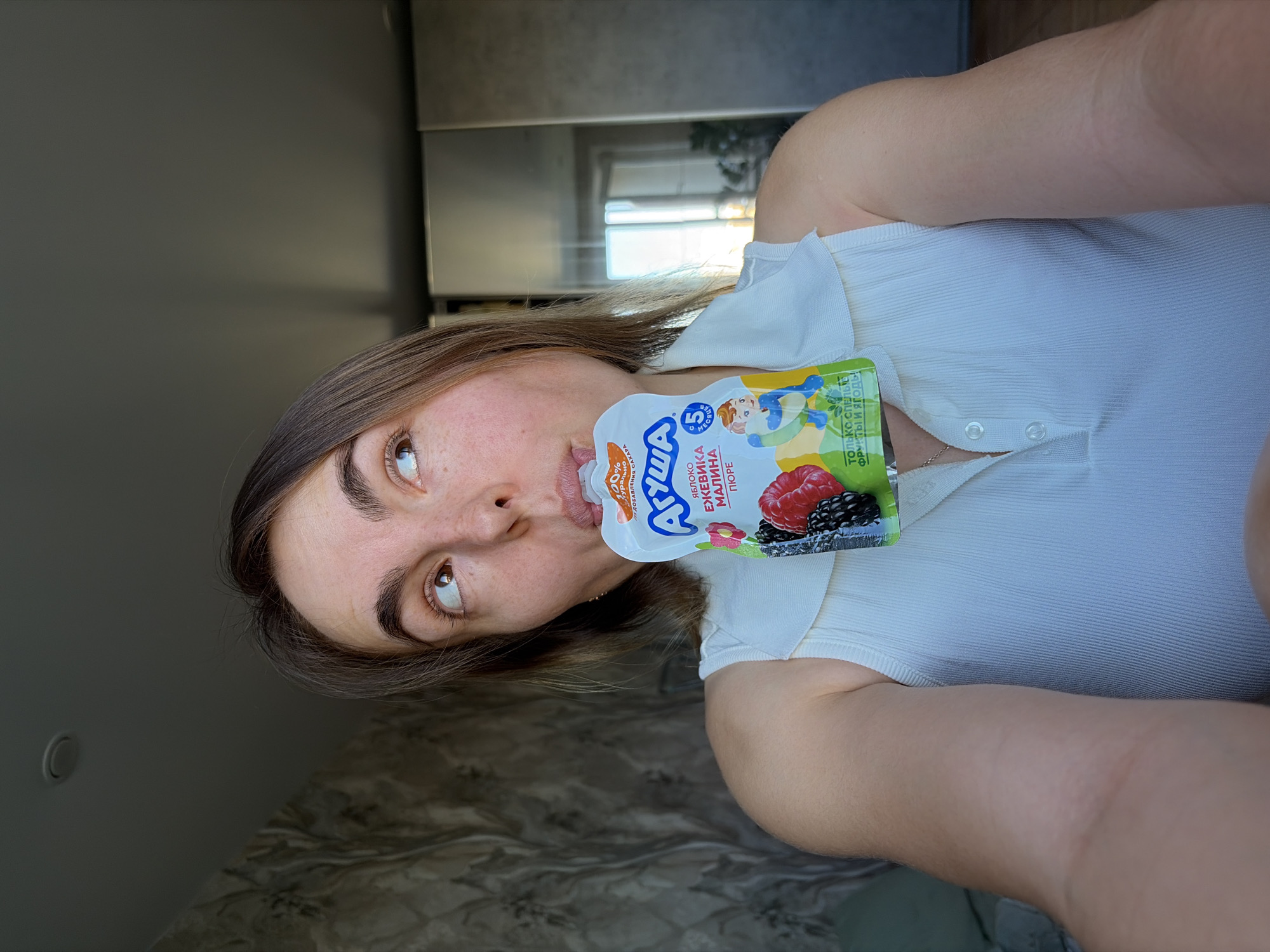 A woman is holding a pouch of "Agusha" fruit puree near her mouth. The puree is labeled "Apple, Blackberry, Raspberry Puree" and is targeted for children ages 5 months and up. The woman is wearing a white top. The background appears to be indoors with furniture and a window visible. The text on the fruit puree is in Russian. It is impossible to know if this is in Izhevsk, Russia.
