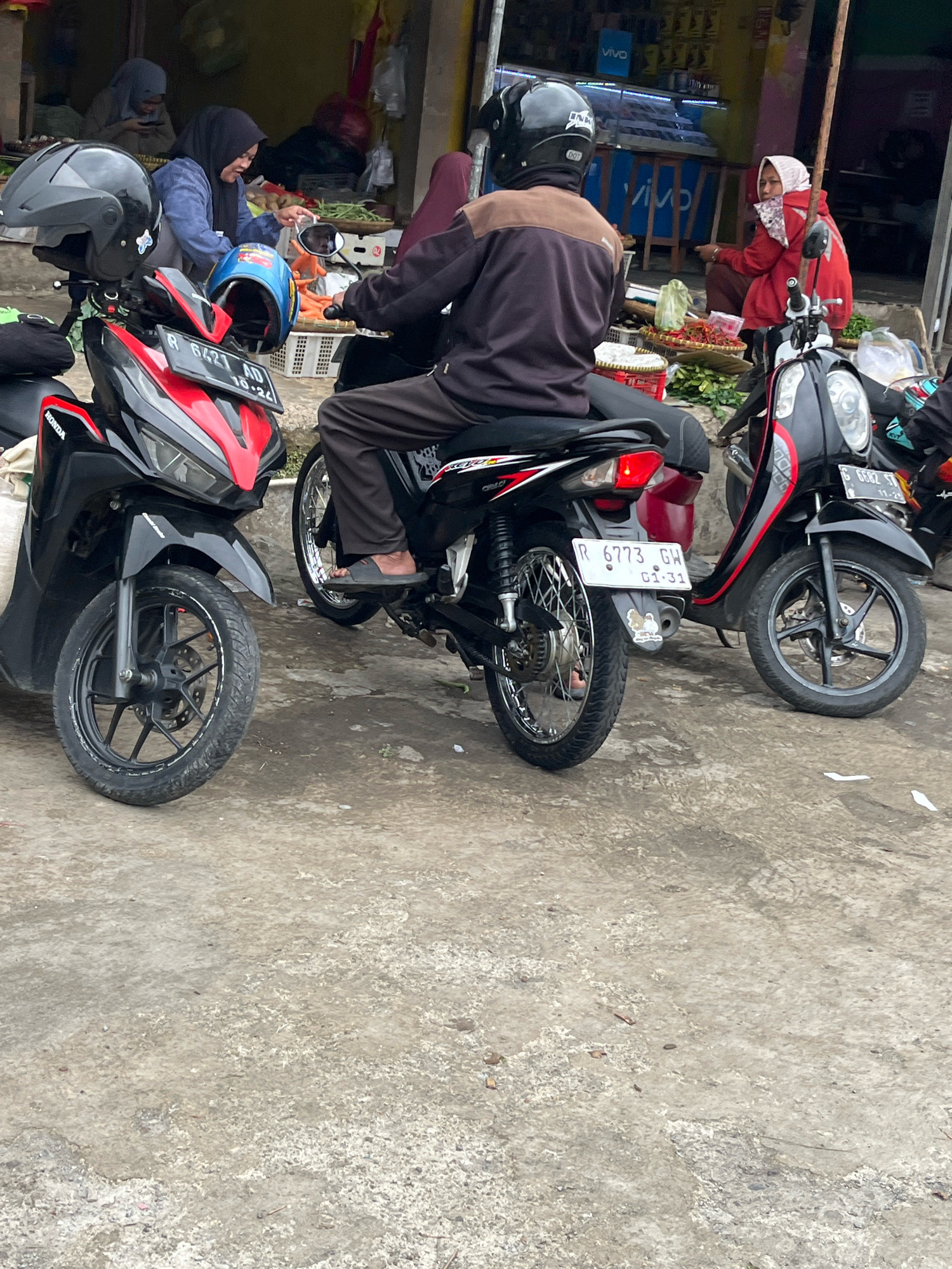 In Karangkobar, Indonesia, three scooters are parked on a concrete surface near market stalls. A person wearing a black helmet and a dark jacket is seated on the middle scooter, which is red and black. To the left, a black and red scooter has two helmets on it, one black and one blue with a cartoon character. Further to the right, a black scooter is parked, and its rider, wearing a red hijab and jacket, is partially visible, seated and looking to the left. Behind the scooters, market stalls are set up with produce, including vegetables and chili peppers, and people in headscarves are visible within the stalls. The environment appears to be an outdoor market with a rustic feel.