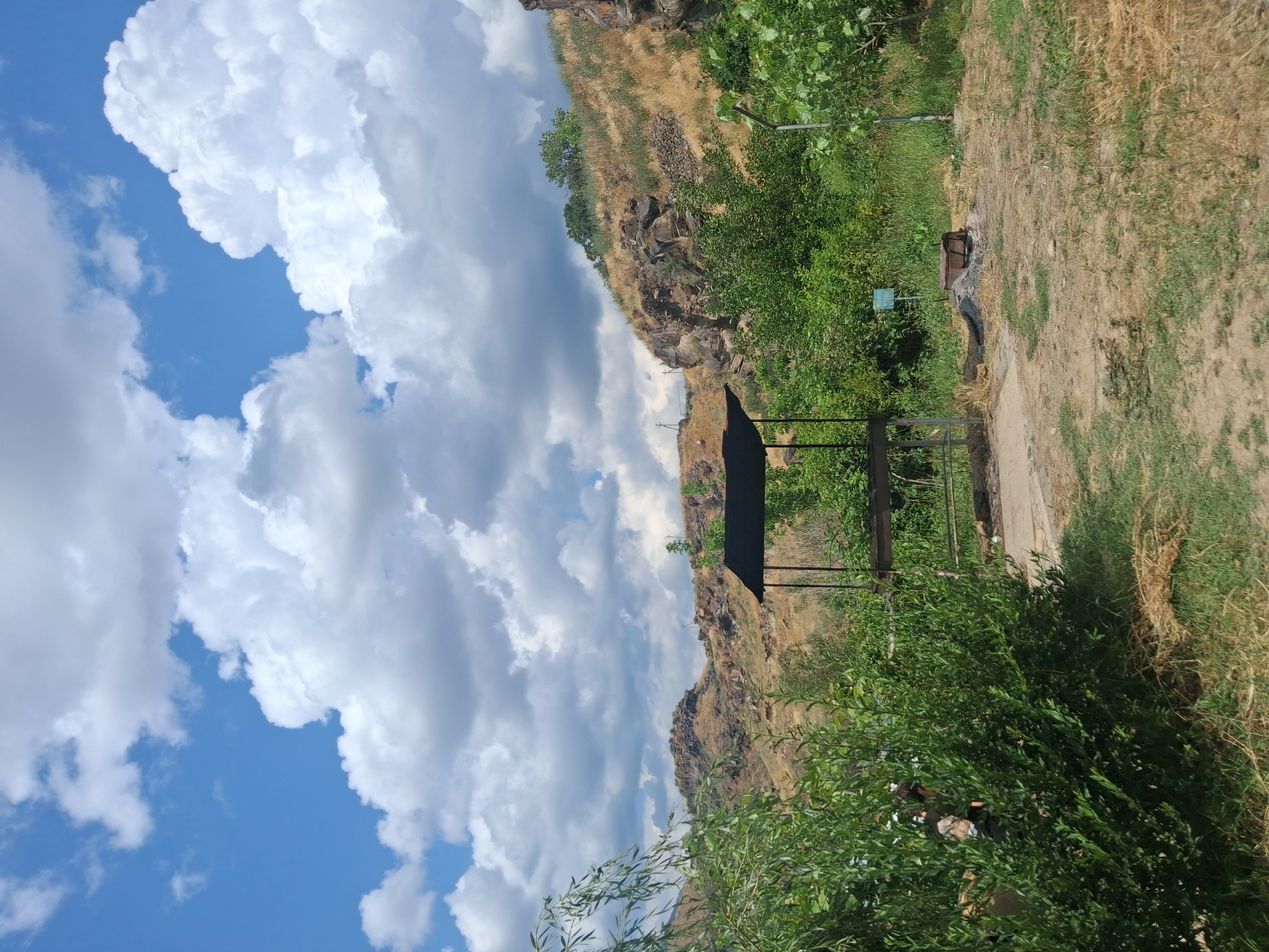 A barbecue area with a metal grill and canopy is situated in a grassy, uneven terrain at the foot of a rocky, dry hillside. The sky above is blue with scattered, puffy white clouds. Lush green vegetation, including trees and bushes, surrounds the area, partially obscuring a person in camouflage clothing. A small, burnt-out fire pit is visible to the right of the grill. The scene appears to be a rustic outdoor recreational spot in Malishka, Armenia.