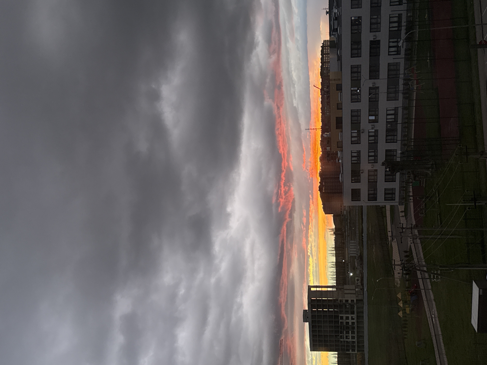 The image depicts a sunset over a city. In the foreground, there are several apartment buildings and green spaces. Power lines and a street with sidewalks are visible. The sky is filled with dark, grey storm clouds on the left side, transitioning into layers of white, orange, and yellow as the sun sets on the right side of the image. The city and location are unknown.