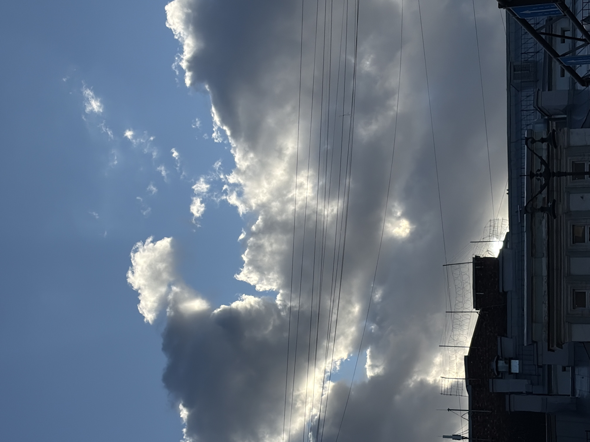 The image depicts a view of the sky above Saint Petersburg, Russia, characterized by dramatic clouds partially obscuring the sun. The sky is a vibrant blue, with large, billowy gray clouds dominating the frame, interspersed with patches of bright sunlight that create rays. Several dark, thin power lines are strung across the middle of the image. Below the sky, the rooftops of buildings are visible, including what appears to be barbed wire or a similar security fencing on top of one of the structures. The architecture suggests an urban environment. No people are present in the frame.