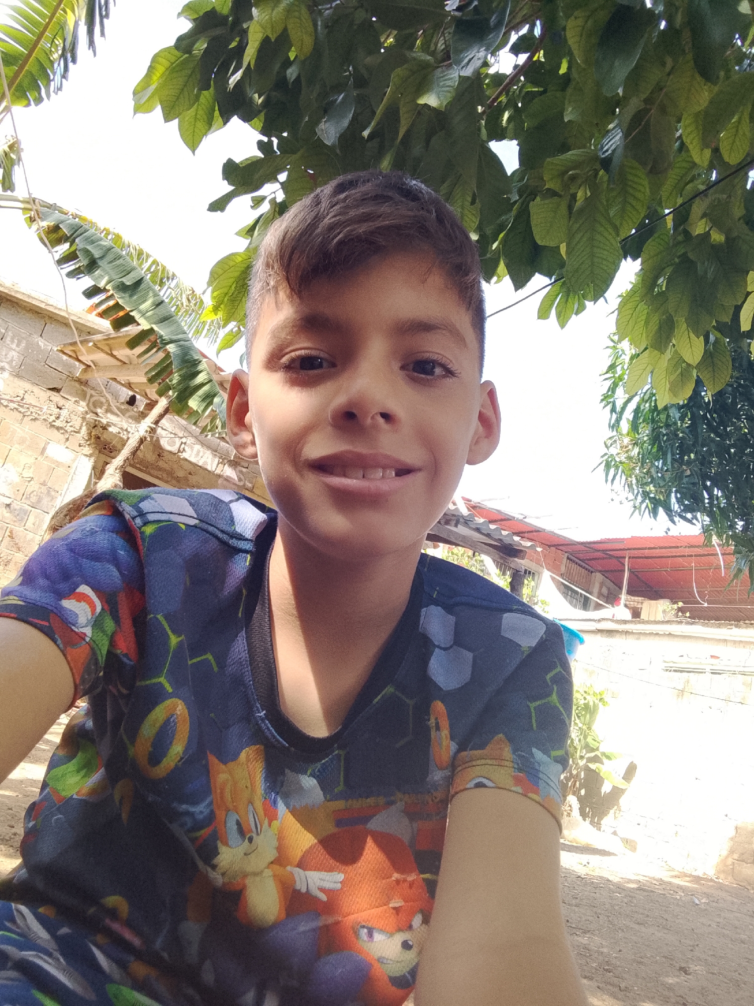 A young boy is taking a selfie outdoors in what appears to be a residential area in Altagracia, Venezuela. He has brown hair, a light complexion, and is wearing a t-shirt with animated characters printed on it. The background shows a partially constructed brick building with a corrugated metal roof, foliage from trees, and a clear sky. The ground appears to be dirt or gravel.