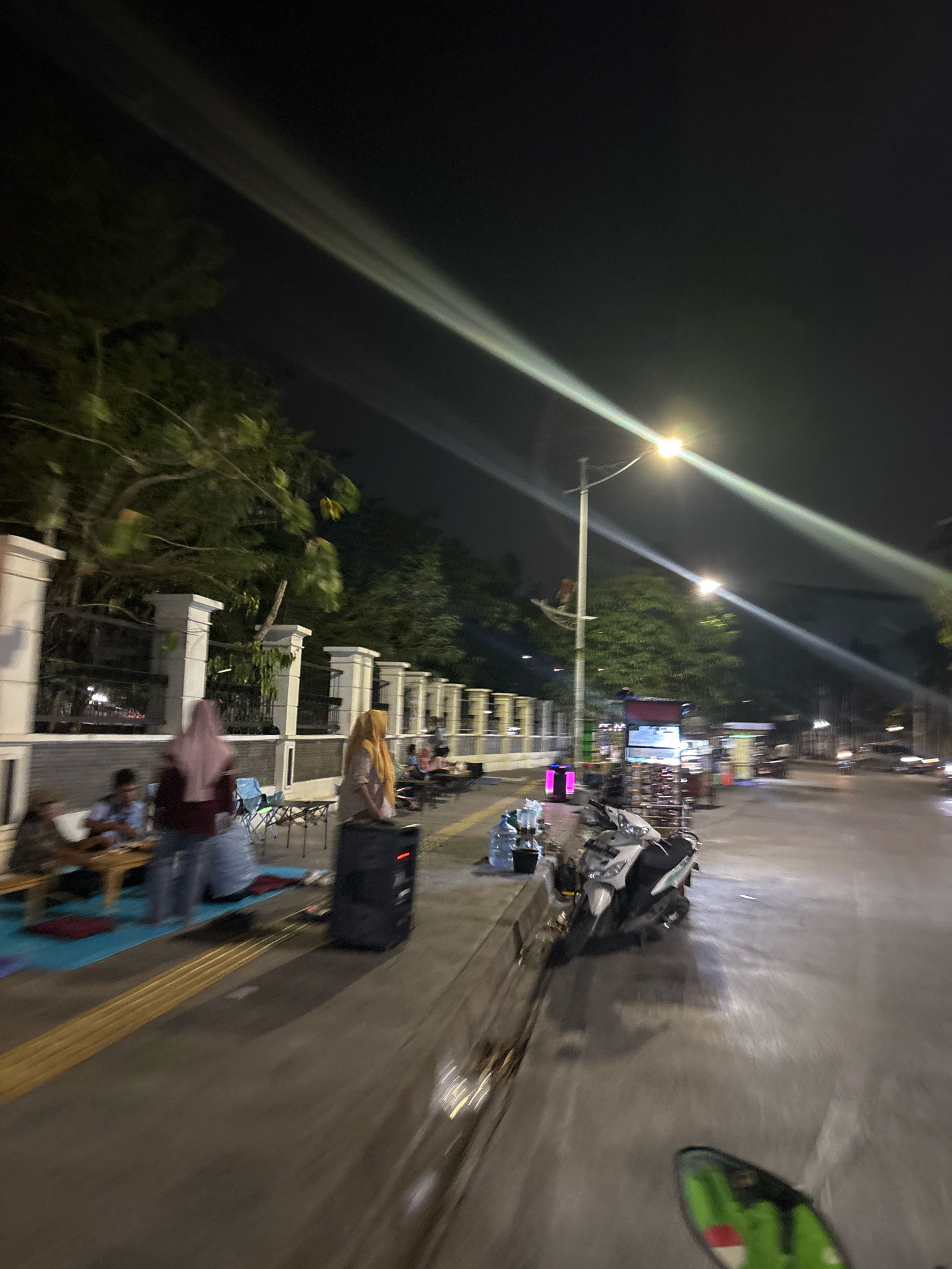 A night scene unfolds on a street in North Jakarta, Indonesia, likely a public walkway adjacent to a road, illuminated by streetlights and the lights from street vendors. In the foreground, a portion of a green and white object, possibly a scooter's handlebar, is visible, suggesting the perspective is from a moving vehicle. To the left, a raised sidewalk area features a white fence with decorative pillars. Several individuals are gathered at low tables, seated on mats and chairs, suggesting they are customers or patrons of a food stall. One person in a maroon top and pink hijab stands near a portable speaker, while another in a yellow hijab stands in front of it. Behind them, a row of street vendors' carts are visible, with some displaying goods under lights. The road to the right shows passing vehicles and more streetlights casting long streaks of light across the scene. Trees line the background, their leaves dark against the night sky. The overall atmosphere is that of a bustling evening street market or gathering place.