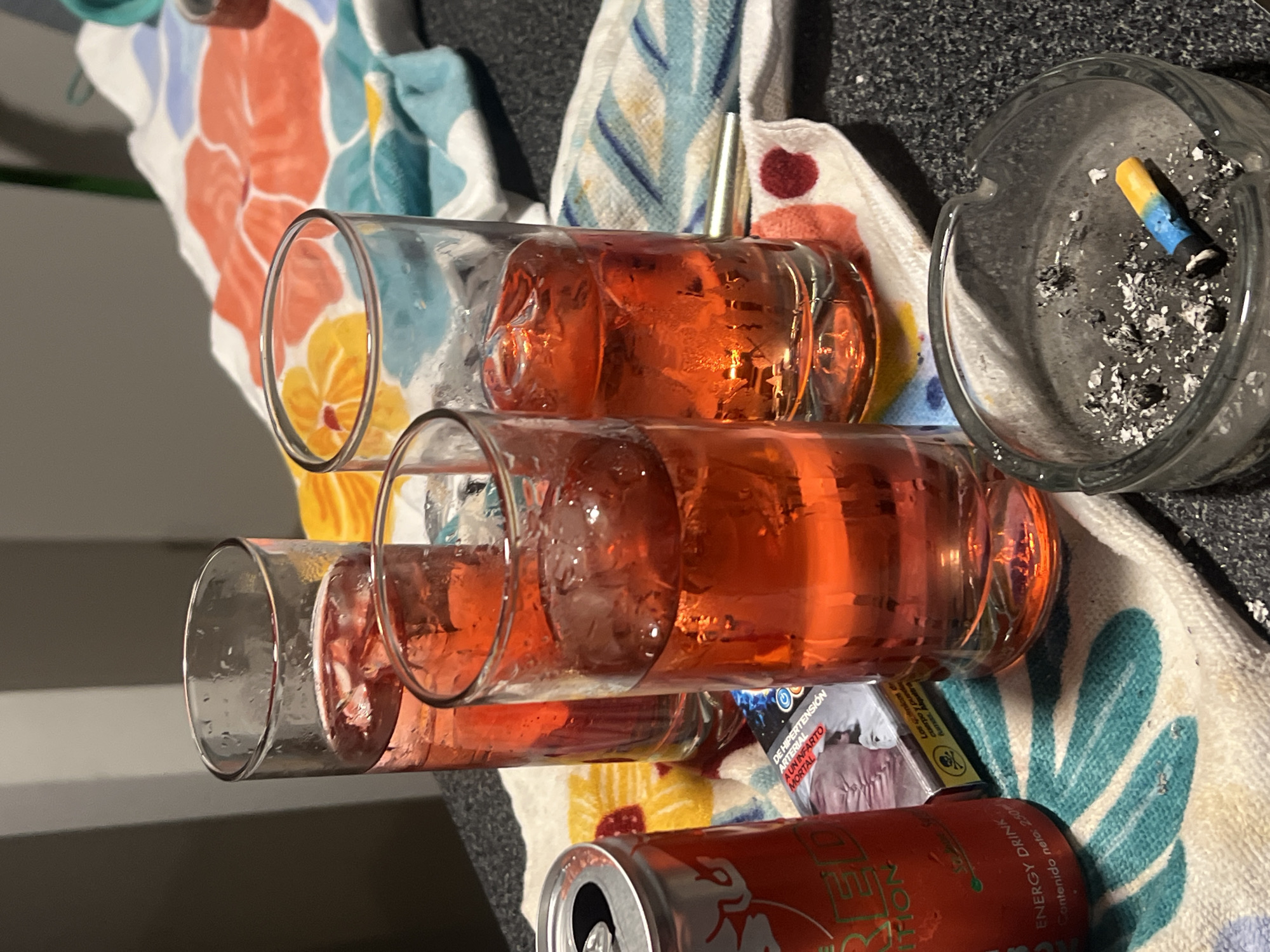The image shows a cluttered scene with various objects on a floral-patterned towel. There are three partially-filled glasses containing a reddish liquid with ice. A red and silver energy drink can is partially visible. A pack of medication with Spanish text is also present. An ashtray containing a cigarette butt and ash is in the upper right corner of the image. The location is likely indoors. The city of Arica, Chile, is not indicated in the image.