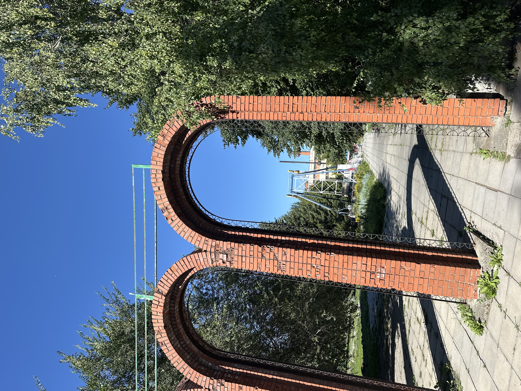 The photograph captures an outdoor scene under a clear blue sky. In the foreground, two arched brick structures with decorative metalwork stand on a paved walkway. Beyond these arches, a path leads towards a distant amusement park ride, characterized by a blue and white structure. Green trees flank the walkway, with one tree prominently visible to the right, its branches extending towards the top of the frame. The ground is covered with a tiled pathway interspersed with patches of grass. The overall impression is that of a sunny day in a park or recreational area.
This image was taken in the city of Biysk, Russia, at the "Lotos" amusement park, located at 22 Vokzalnaya Street.