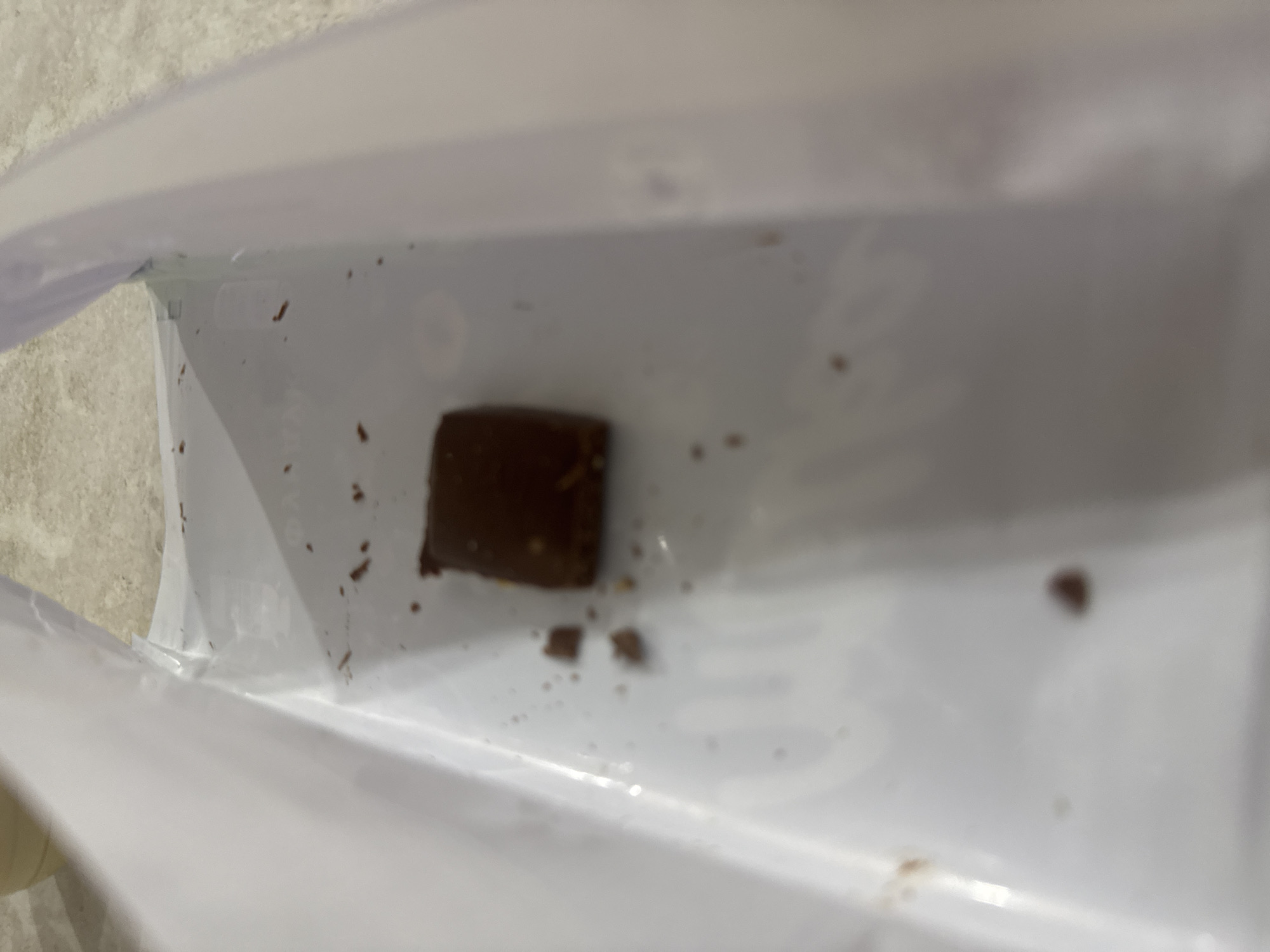 A single, small, square piece of dark chocolate rests on a white surface. The chocolate appears to be slightly melted or smudged. Scattered around the chocolate are a few smaller crumbs and specks of chocolate debris. The white surface seems to be the interior of a wrapper or container, possibly made of plastic or foil. The background is out of focus and appears to be a countertop with a tiled pattern. There are no people or any other identifying elements in the image to suggest a specific location, including Saratov, Russia.