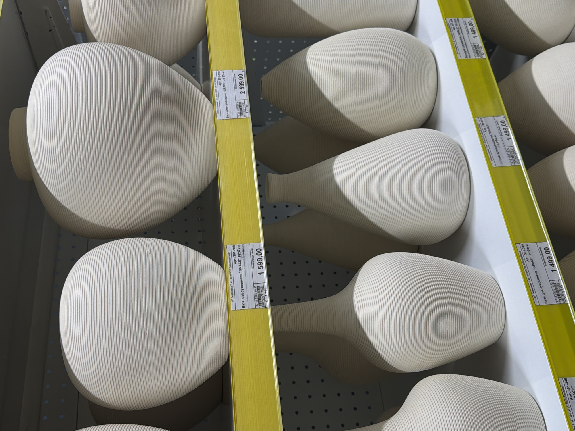 The image shows multiple shelves in a retail environment, filled with ceramic vases of various sizes and shapes. The vases are primarily light beige in color and feature a ridged texture. Yellow price tags are attached to the shelving, indicating the prices of the vases, ranging from 1499.00 to 2599.00. The price tags also include identifying information such as "Арт. 391-346" and the description "Ваза для сухоцветов 'GRACE'," along with dimensions in centimeters (e.g., "14*30см"). The tags further identify the vendor as "ИП Гимадеева Т. И." and provide their INN/КПП numbers, which indicate that the location is within Russia, specifically Perm (as inferred from the INN number). The labels also show dates and times (e.g., "17.11.2025 19:52") likely indicating when the prices were last updated or when the items were placed on the shelf. The shelves are light gray, and the lighting appears to be fluorescent.