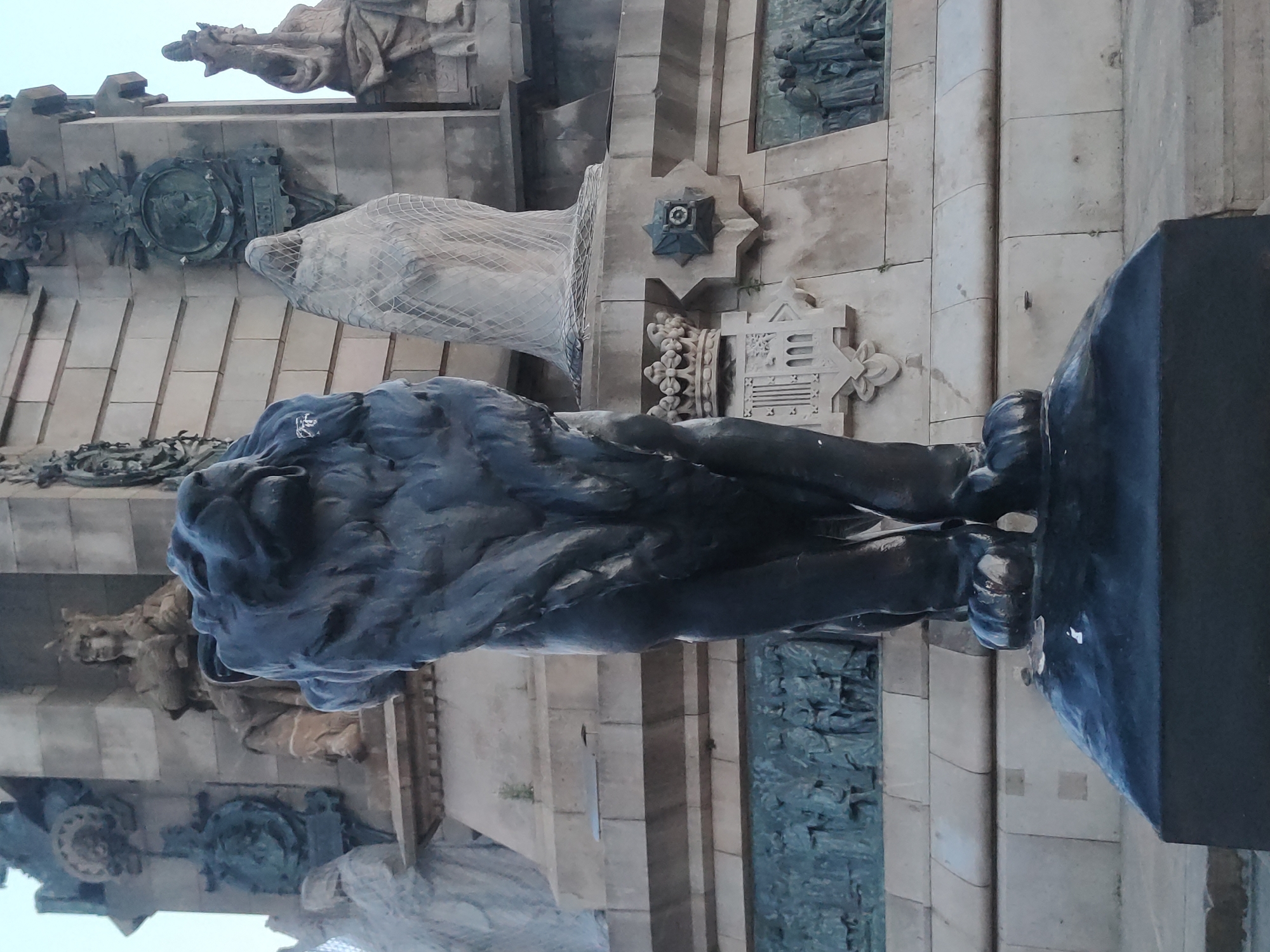 A low-angle, eye-level medium shot captures a dark, imposing statue of a lion standing guard in front of a large, ornate monument in Barcelona, Spain. The lion, sculpted from dark stone or metal, is positioned in the foreground, facing slightly left and looking upwards with an open mouth and powerful physique. Its body is detailed, showing musculature and its paws are firmly planted on a dark, rectangular pedestal.
Behind the lion, the monument rises, constructed from light-colored stone blocks. The monument features numerous sculptures and reliefs. To the left of the lion, a partially visible veiled figure, seemingly made of stone, is wrapped in netting. Further back, a seated figure wearing a crown is depicted in a niche. To the right of the veiled figure and directly behind the lion, a coat of arms with a crown on top is visible, flanked by decorative elements. Higher up on the monument are round medallions and other sculpted figures, some in niches. The background is a light, overcast sky.
The overall impression is one of grandeur and historical significance, with the lion acting as a sentinel to the monumental structure. The monument is likely the Monument a Colom (Columbus Monument) in Barcelona, given its iconic lion statues and proximity to the waterfront.