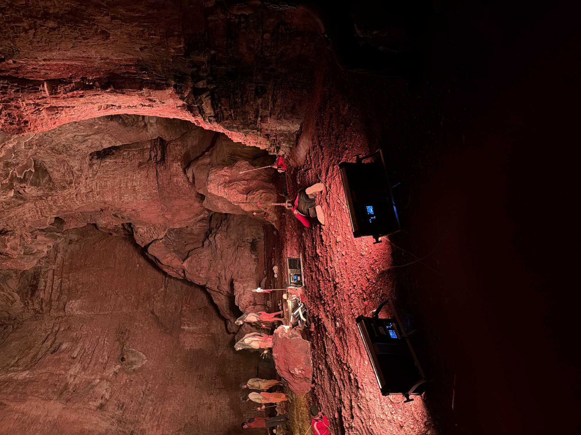 The image depicts an indoor scene within a cave illuminated by red lighting. In the background, several individuals dressed in orange garments and one person in a red jacket are standing or observing. Some figures are positioned near a large rock. In the middle ground, a person is lying face down on the ground, with a red rope attached to their body. Several pieces of equipment, including what appear to be lights and a control panel, are placed on the red, gravelly floor. The environment is characterized by rough, textured cave walls and a floor covered in red gravel. The scene appears to be a staged or artistic setup, possibly for a performance or photoshoot, given the lighting and props. The specific location in Guttapadu, India, is not visually evident from the provided image.