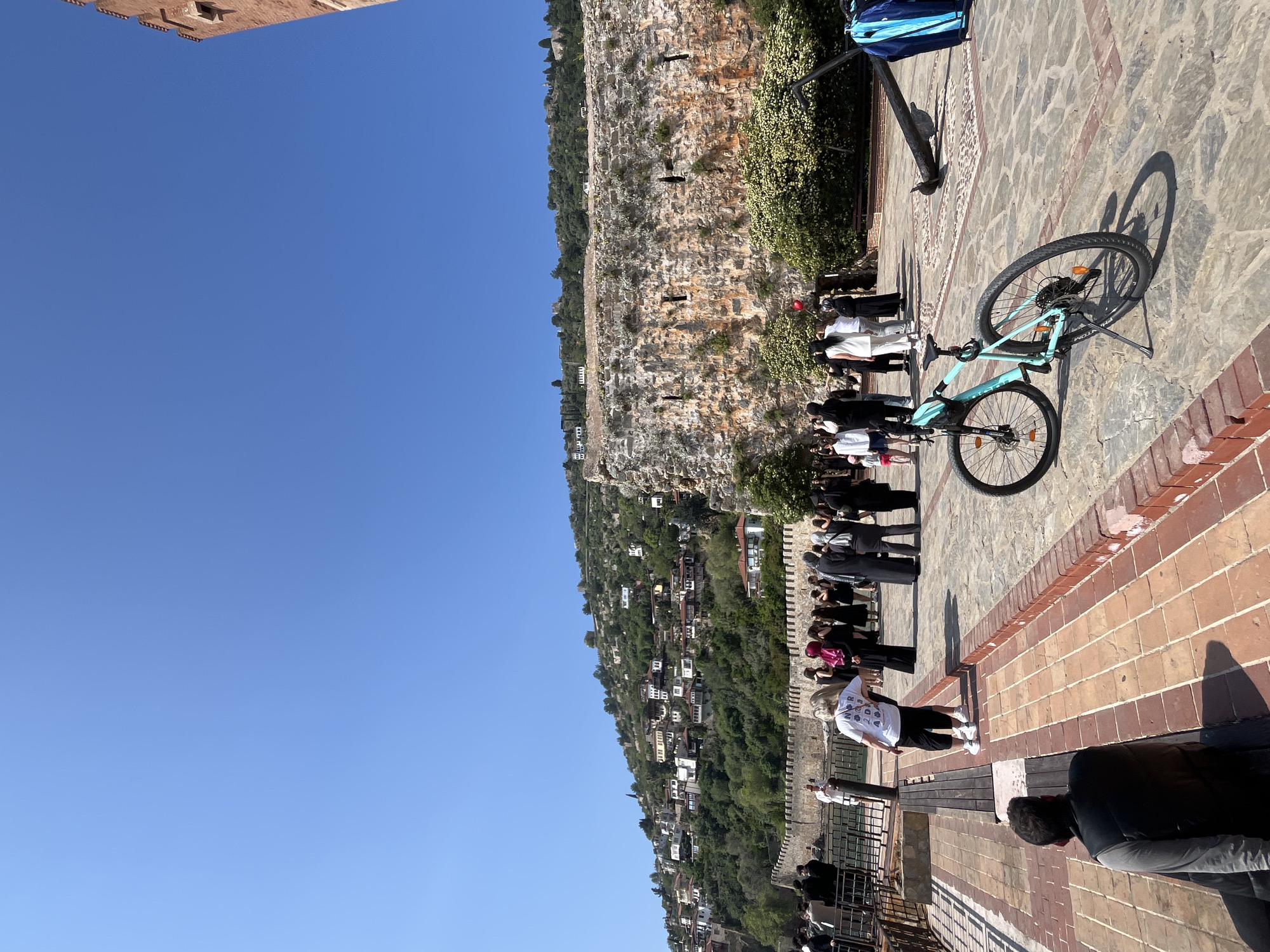 This image, taken from a scenic overlook in Alanya, Turkey, depicts a bright, sunny day with people observing a panoramic view. In the immediate foreground, a light blue "SCOTT" brand mountain bike is parked on a patterned stone pavement, casting a clear shadow. A low brick wall outlines part of the paved area.
A group of people are gathered in the middle ground, standing along a stone wall and looking out towards the distant scenery. Their clothing varies, with many wearing dark attire, while a woman in a white t-shirt and black shorts stands closer to the foreground. Another individual, partially visible in the lower right, also faces the view. A large blue and white bag is visible in the upper right corner, near an iron post.
The background showcases a dramatic landscape under a vast, clear blue sky. A prominent, rugged rock face or cliff, interspersed with sparse vegetation, dominates the left side of the frame. Below this, a lush, green hillside descends, densely dotted with numerous small, light-colored buildings, characteristic of a hillside town. The bright sunlight and distinct shadows suggest a time between mid-morning and early afternoon. A light brown stone structure is partially visible in the top left, indicating the elevated position from which the photo was taken. The scene conveys a sense of peaceful sightseeing and appreciation for the beautiful natural and architectural setting.