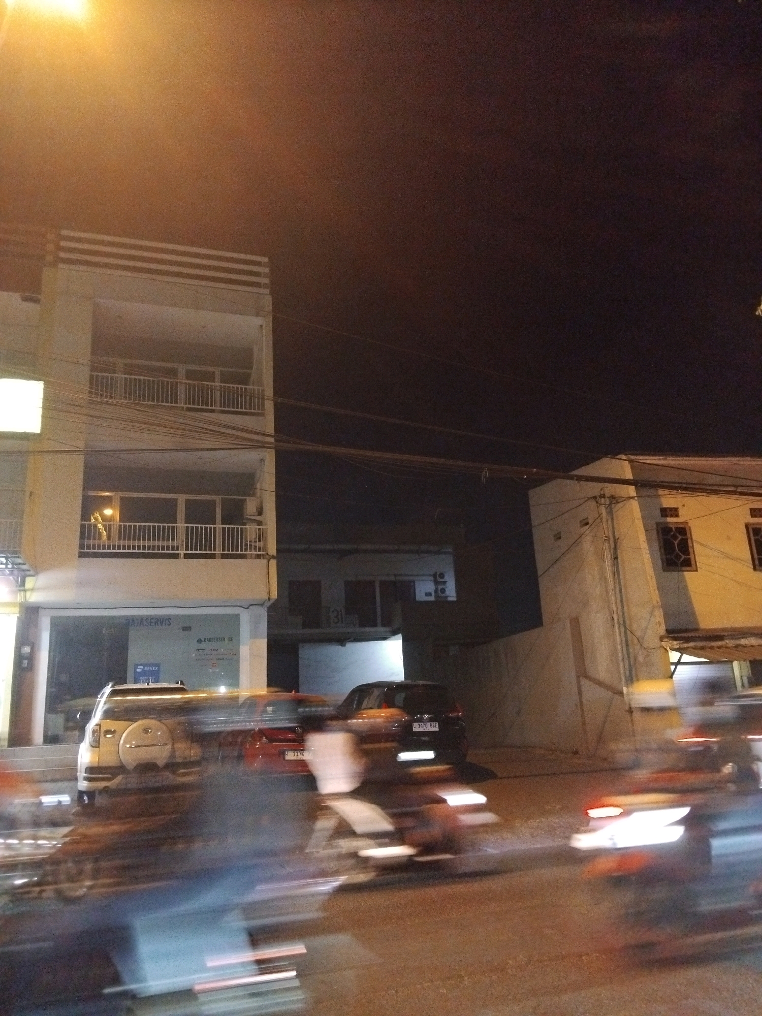 The image captures a nighttime scene on a city street in Wedoro, Indonesia. The focus is on a multi-story building with a balcony on the second floor and a store or office on the ground floor, identified by signage that includes "RAJASERVIS" and "BADBER SERVICE". Adjacent to this building, another structure is visible with windows and a sign displaying the number "31". The street in front of the buildings is busy with motion blur, indicating moving vehicles and possibly pedestrians. Several cars are present, including a white SUV parked in front of the building and other vehicles in motion. The overall lighting suggests streetlights and building illumination, with a warm, orange glow emanating from an overhead light source at the top left of the frame. The sky is dark, typical of nighttime.